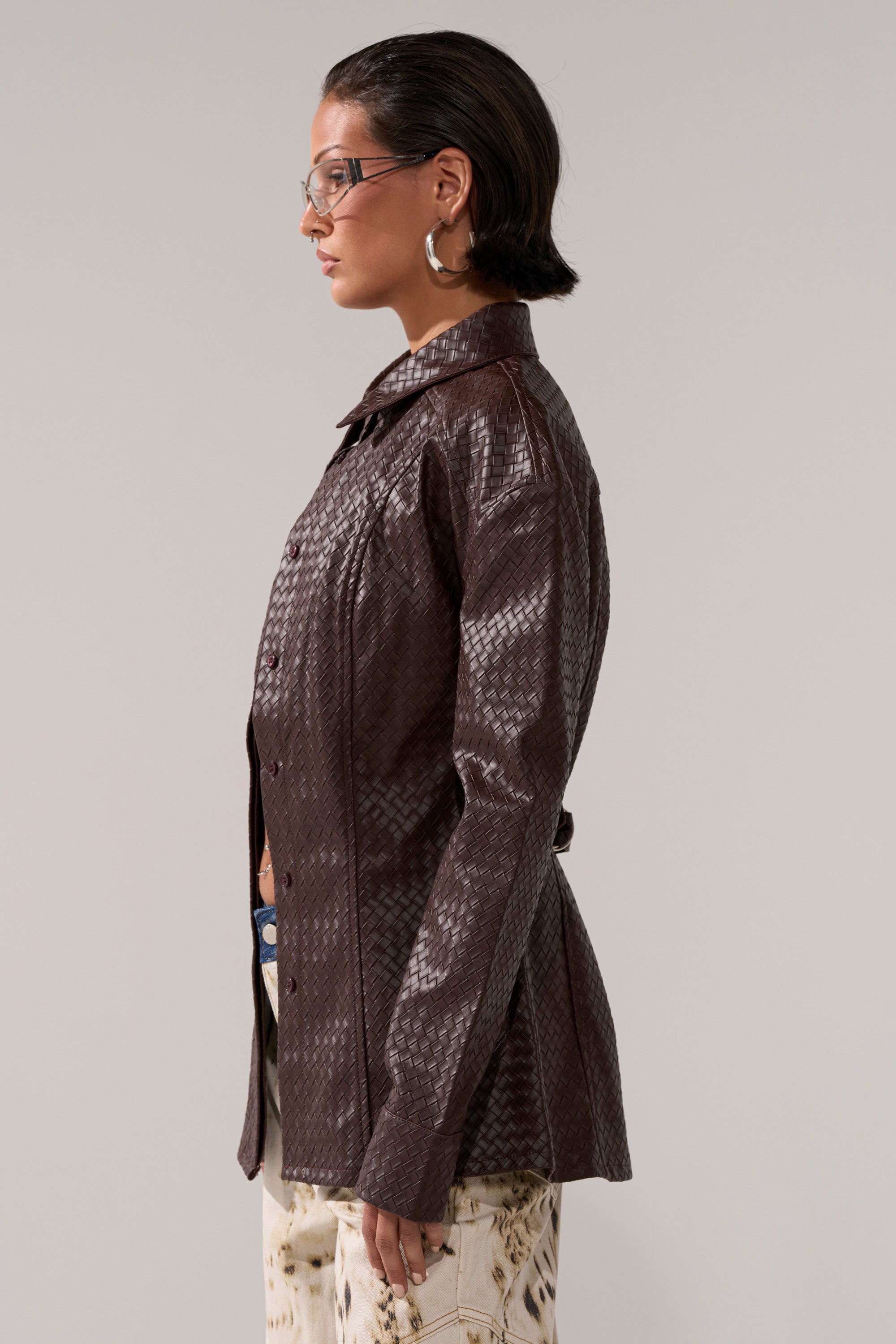 A person with short dark hair, glasses, and hoop earrings models the FEELING RICH FAUX LEATHER BUTTON DOWN—a brown, textured jacket—paired with light patterned pants, standing in profile against a plain background.
