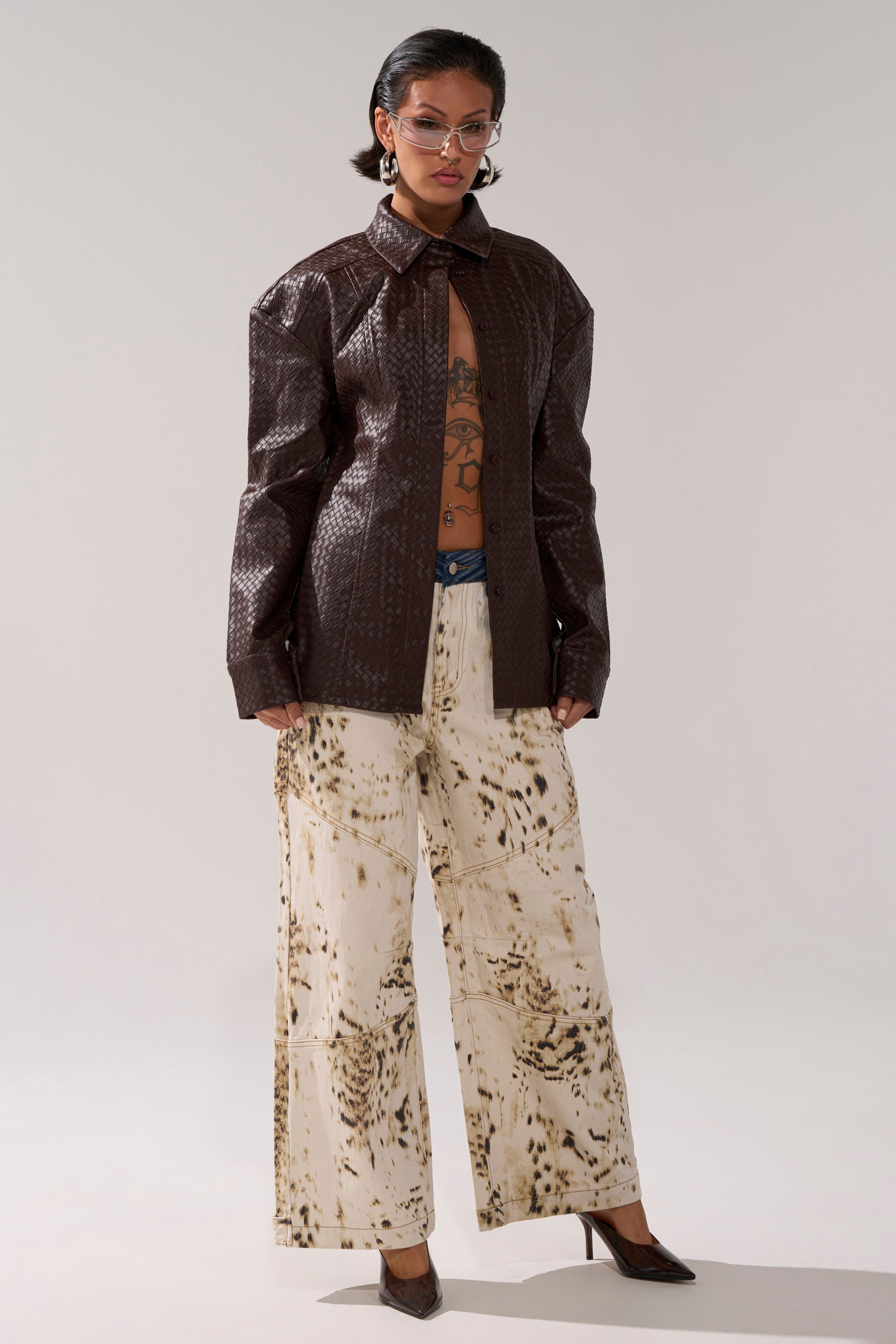 A person stands confidently in the FEELING RICH FAUX LEATHER BUTTON DOWN jacket, paired with cream and brown patterned wide-leg pants, pointy heels, glasses, large earrings, visible chest tattoos, and slicked-back hair.