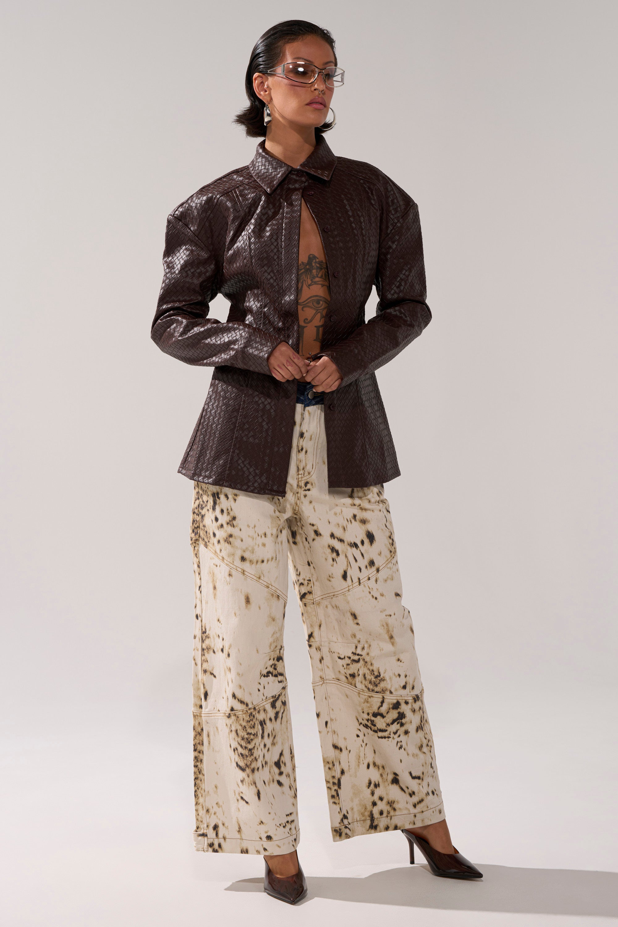 A person wears the FEELING RICH FAUX LEATHER BUTTON DOWN—a textured dark brown jacket with a deep neckline and adjustable buckled belt—paired with wide-leg abstract print pants, brown heels, clear glasses, and displays chest tattoos against a plain background.