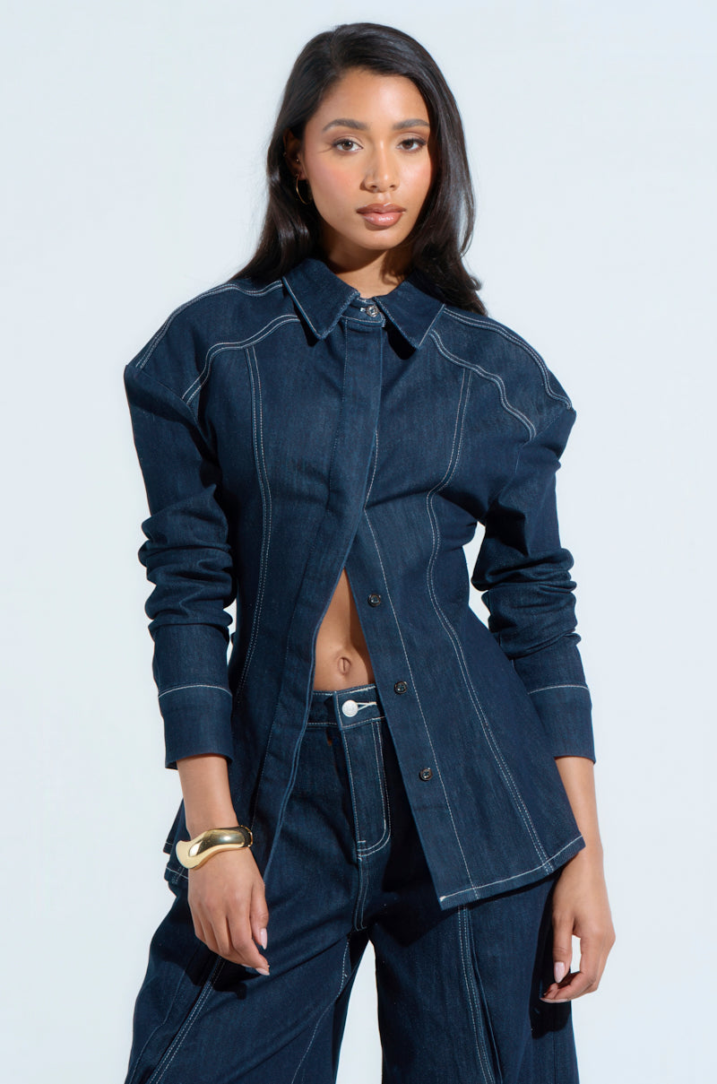 A person in a COME WITH ME BUTTON DOWN TOP with an oversized, relaxed fit denim jacket and matching pants featuring white stitching poses against a plain background, displaying long dark hair and wearing a gold bracelet on their left wrist.