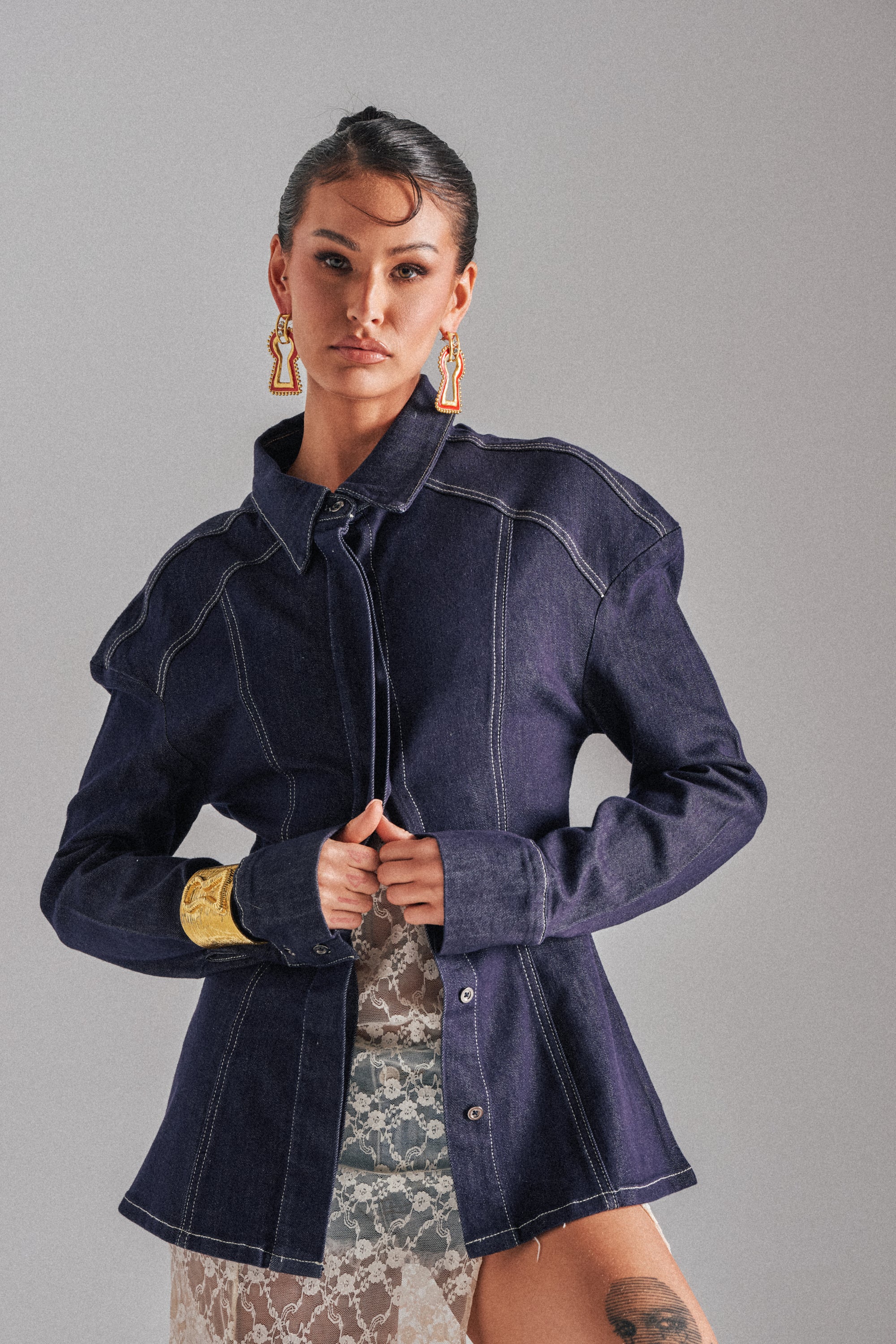 A woman stands confidently against a plain background wearing the COME WITH ME BUTTON DOWN TOP, featuring dark denim, paneled construction, white stitching, gold earrings, a chunky gold bracelet, and a sheer white lace skirt. 0