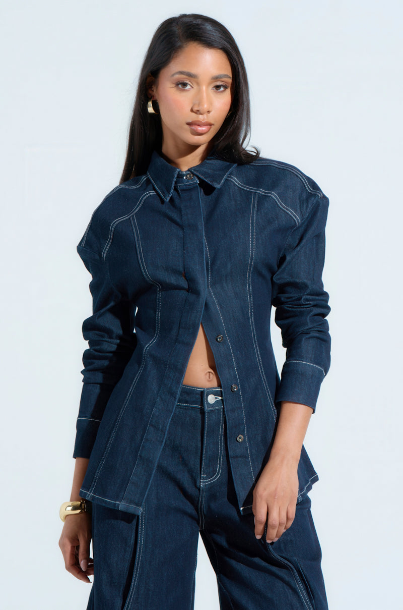 Wearing the "COME WITH ME BUTTON DOWN TOP," a woman in a blue denim jacket and matching Bermuda shorts poses against a light background. The oversized relaxed silhouette complements her long dark hair. Accessorized with a gold bracelet and earrings, her hands rest at her sides.