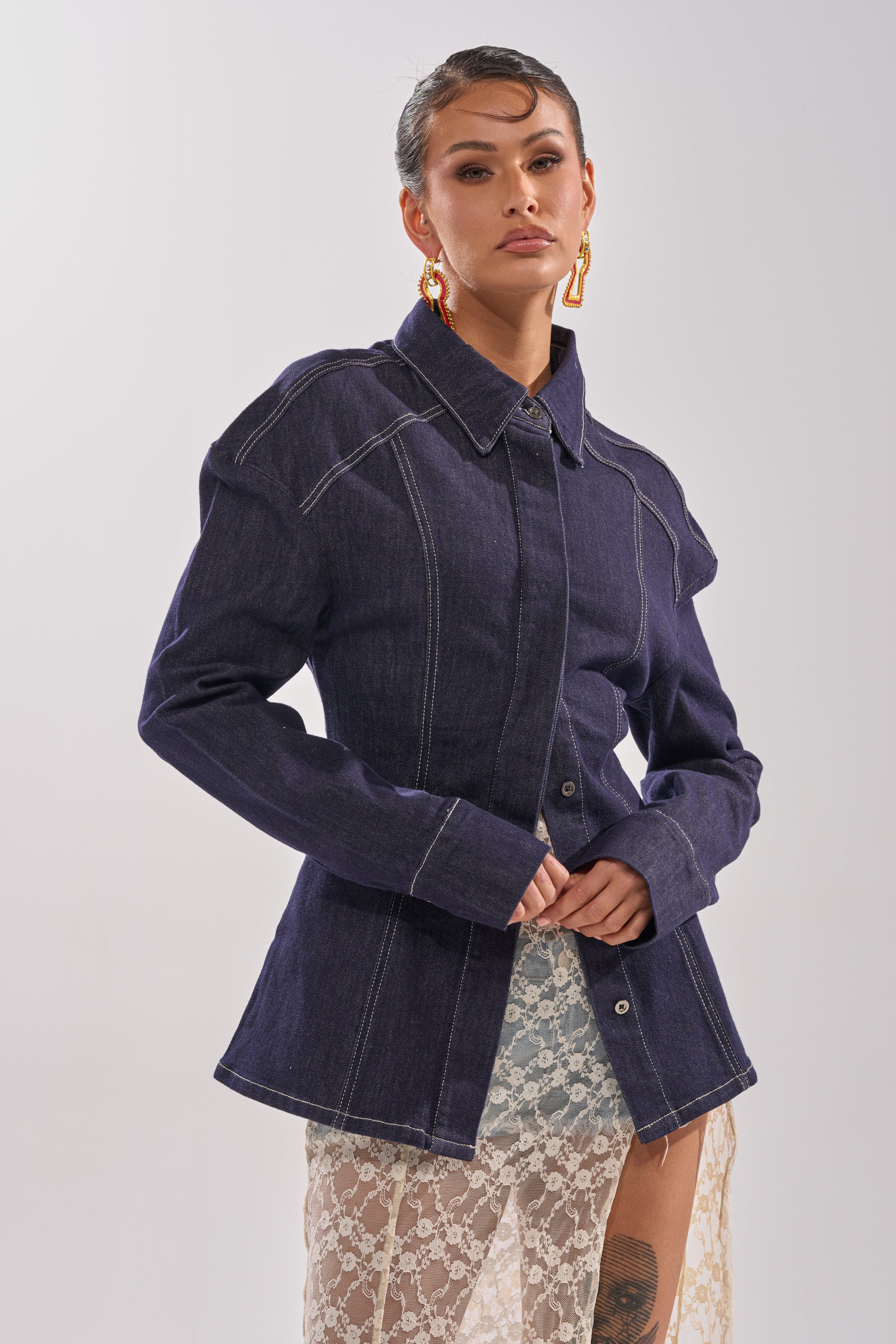 A person wears the COME WITH ME BUTTON DOWN TOP—an AKIRA Label oversized denim shirt with white stitching—paired with large gold earrings and a sheer lace skirt, standing against a plain light backdrop and gazing confidently at the camera. 3
