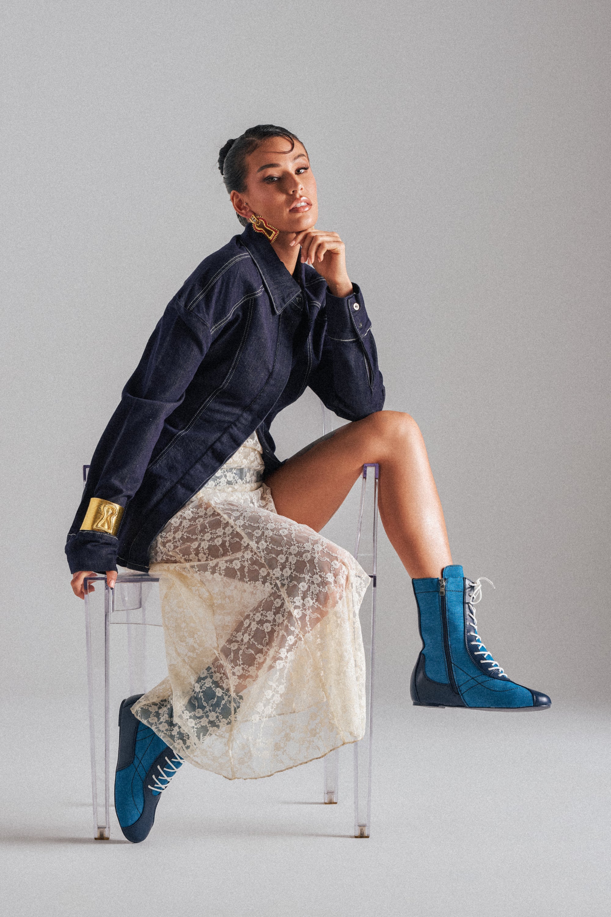 A person sits on a transparent chair, wearing a sheer lace dress under the COME WITH ME BUTTON DOWN TOP, paired with bright blue lace-up boots. One hand rests on their chin as they look confidently at the camera. 1
