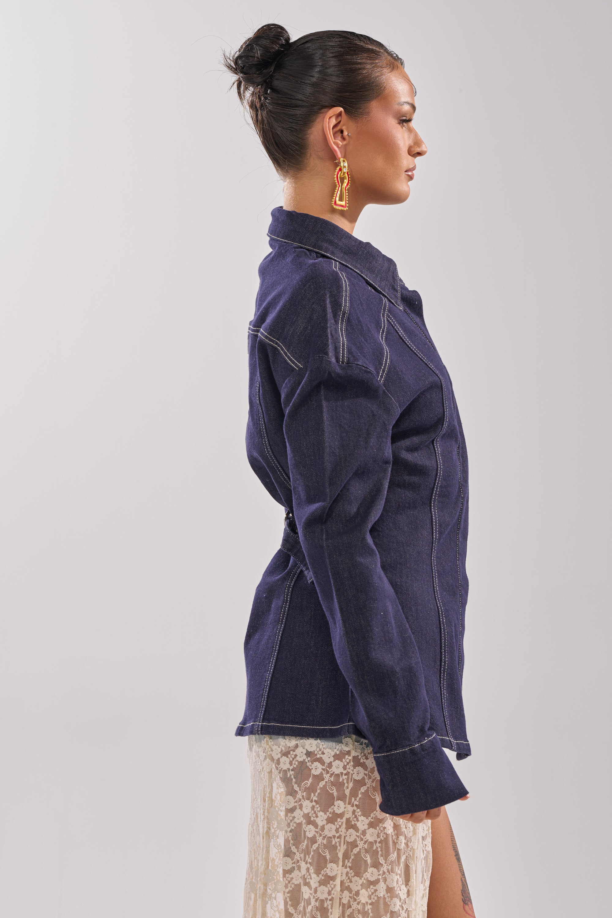 A woman with dark hair in a bun, wearing orange abstract earrings, the COME WITH ME BUTTON DOWN TOP by AKIRA Label, and a cream lace skirt stands in profile against a plain light background. 4