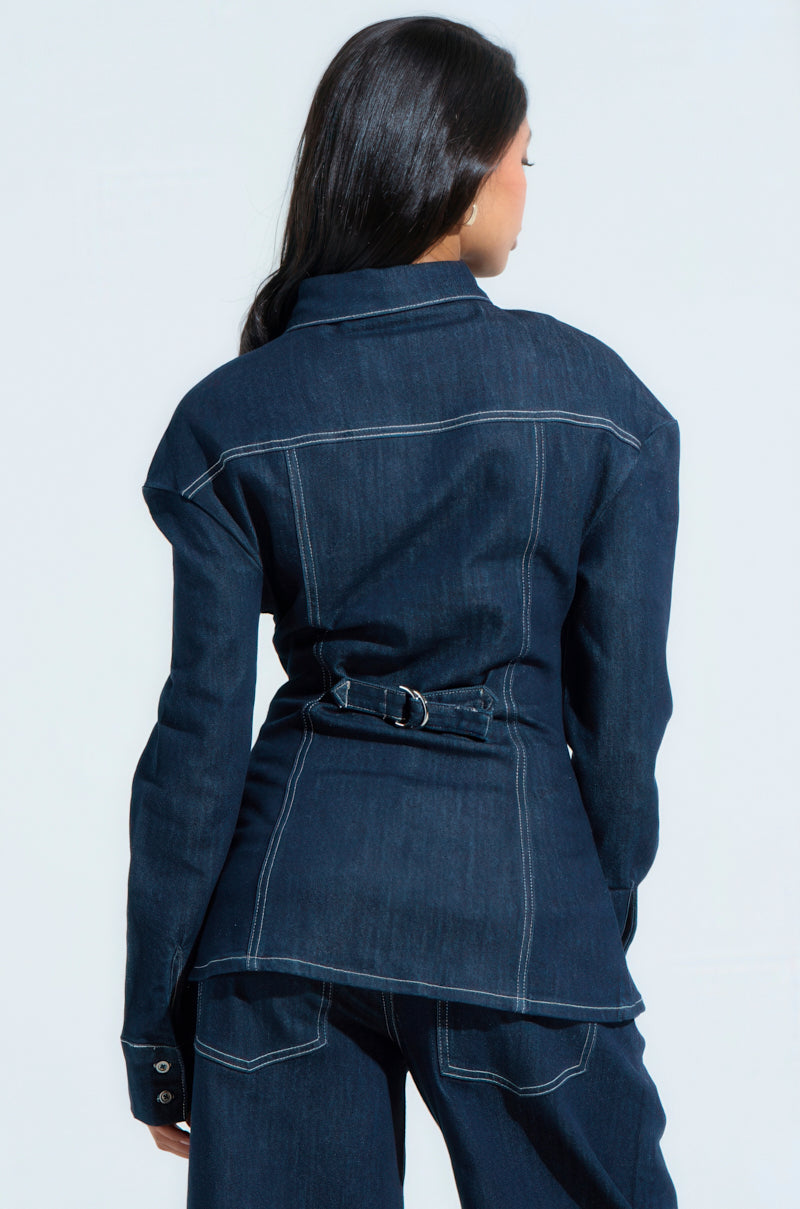 A person with long hair is seen from the back, wearing an oversized COME WITH ME BUTTON DOWN TOP in dark denim with matching Bermuda shorts. The top features white stitching and a belt detail at the waist, set against a light, neutral background that highlights the casual yet stylish look.