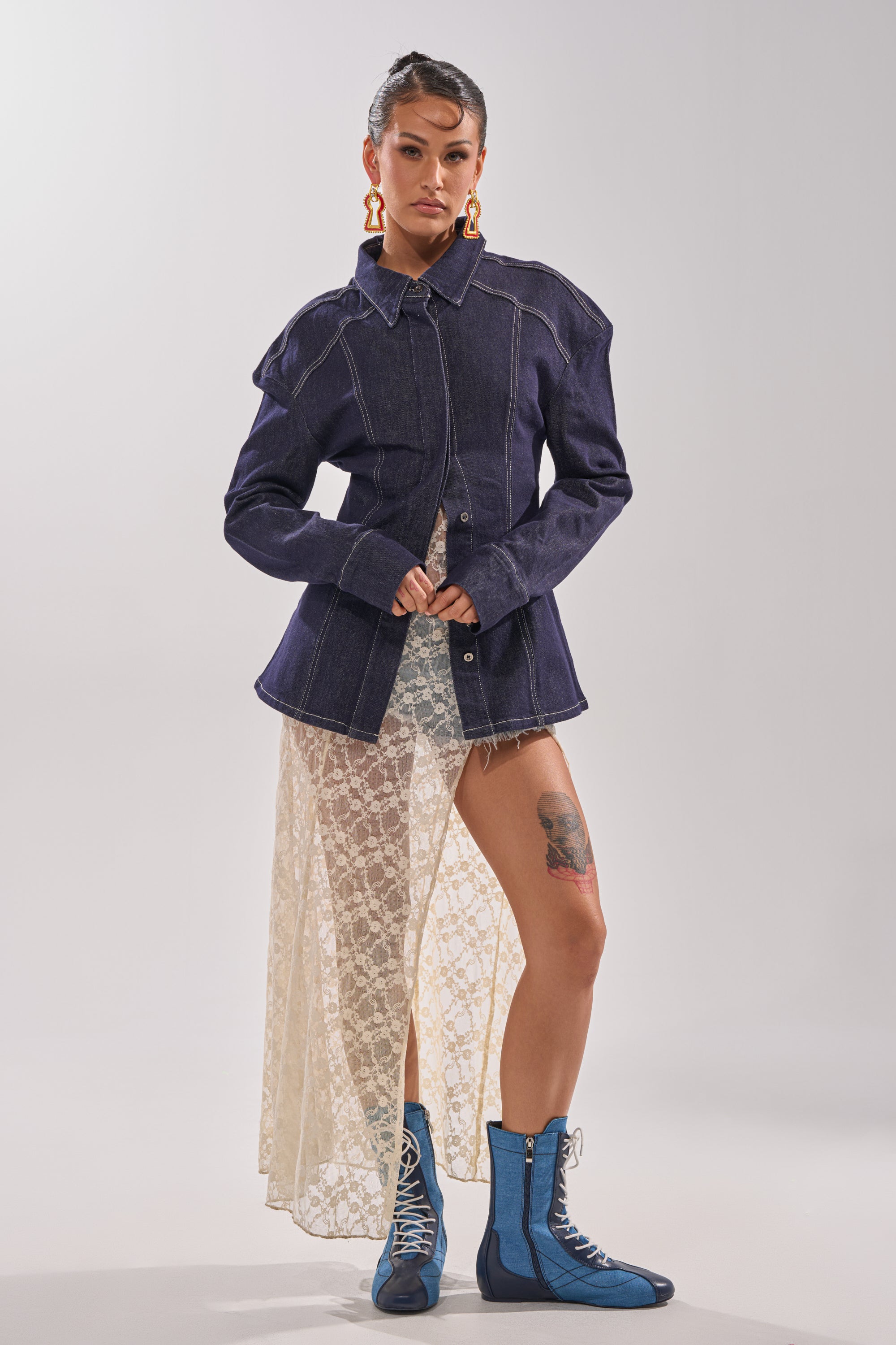 A person stands confidently in the COME WITH ME BUTTON DOWN TOP, paired with a lace skirt, blue lace-up boots, statement earrings, and reveals a thigh tattoo against a plain background. 6