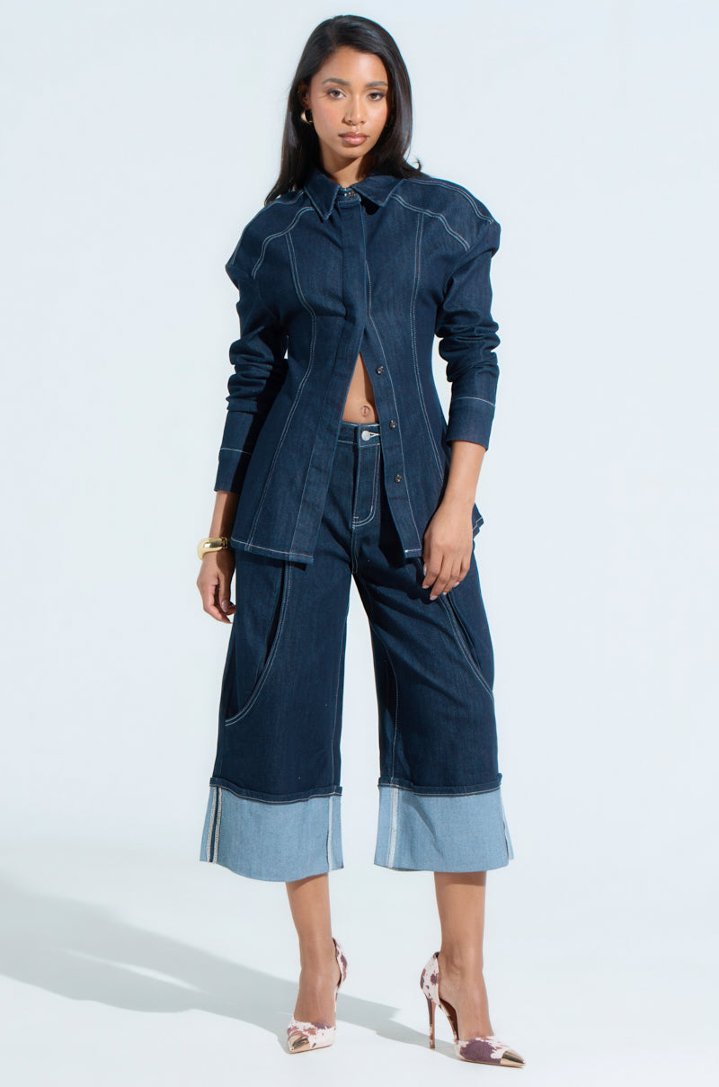 A woman wears the oversized COME WITH ME BUTTON DOWN TOP and wide-legged cropped jeans with lighter cuffs. She completes her look with floral-patterned heels, standing poised against a plain light background.