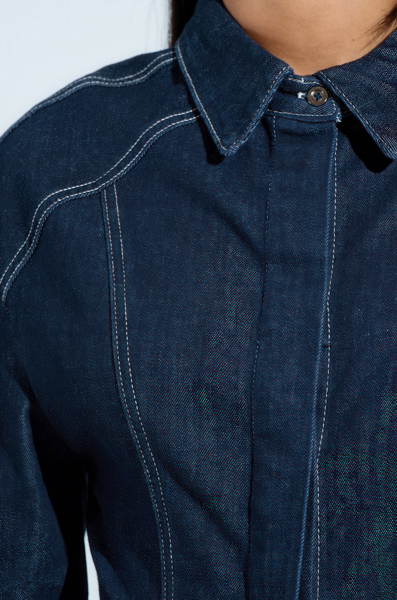 A person wears the COME WITH ME BUTTON DOWN TOP, a dark blue cotton blend denim shirt with white stitching. It features an oversized relaxed fit, a button-up collar, and visible seams against a pale backdrop.