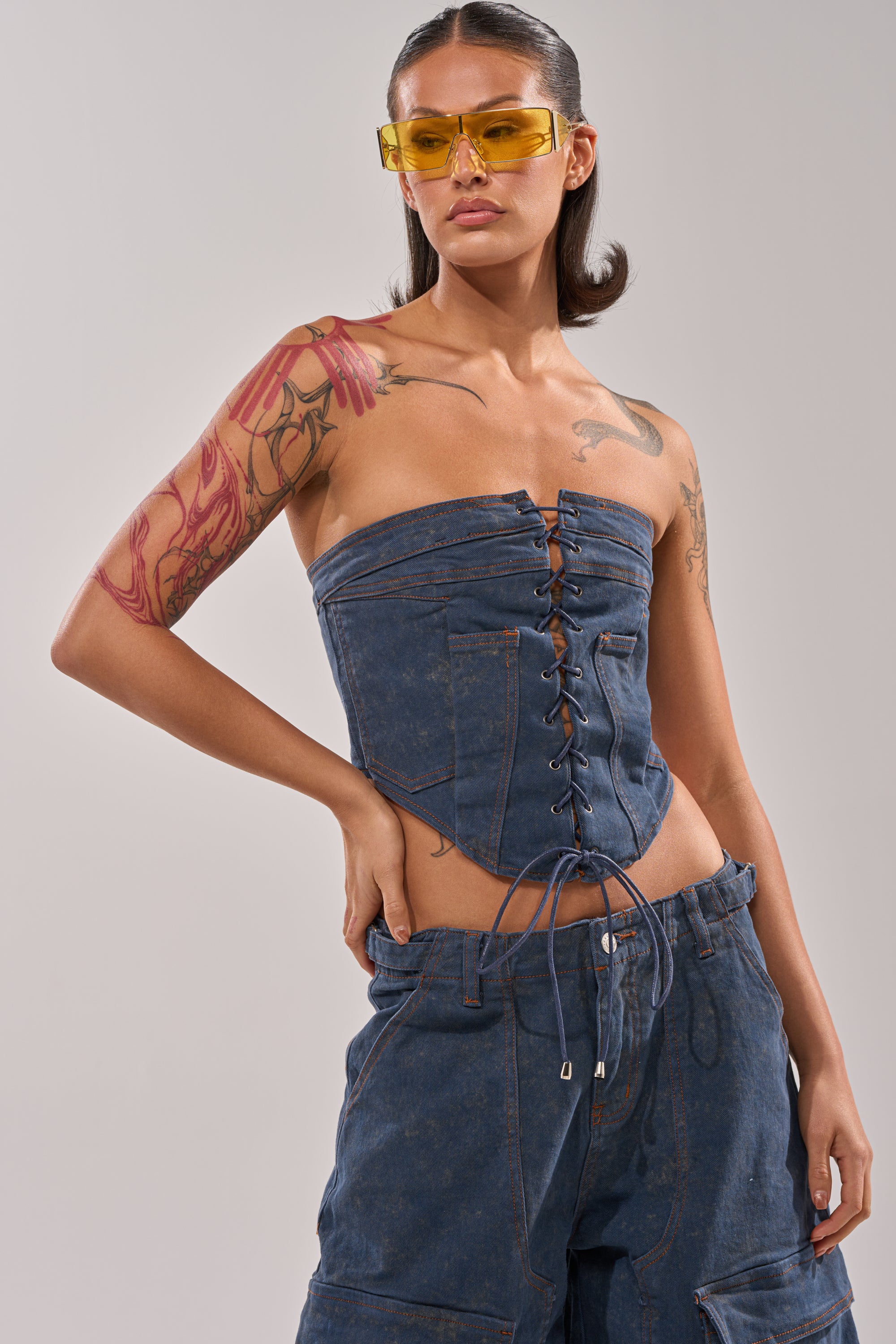 Wearing yellow sunglasses and the BOLD LACE UP CORSET with matching denim pants, this person stands confidently with one hand on their hip. Their straight, dark hair is styled back and tattoos decorate their arms. 0