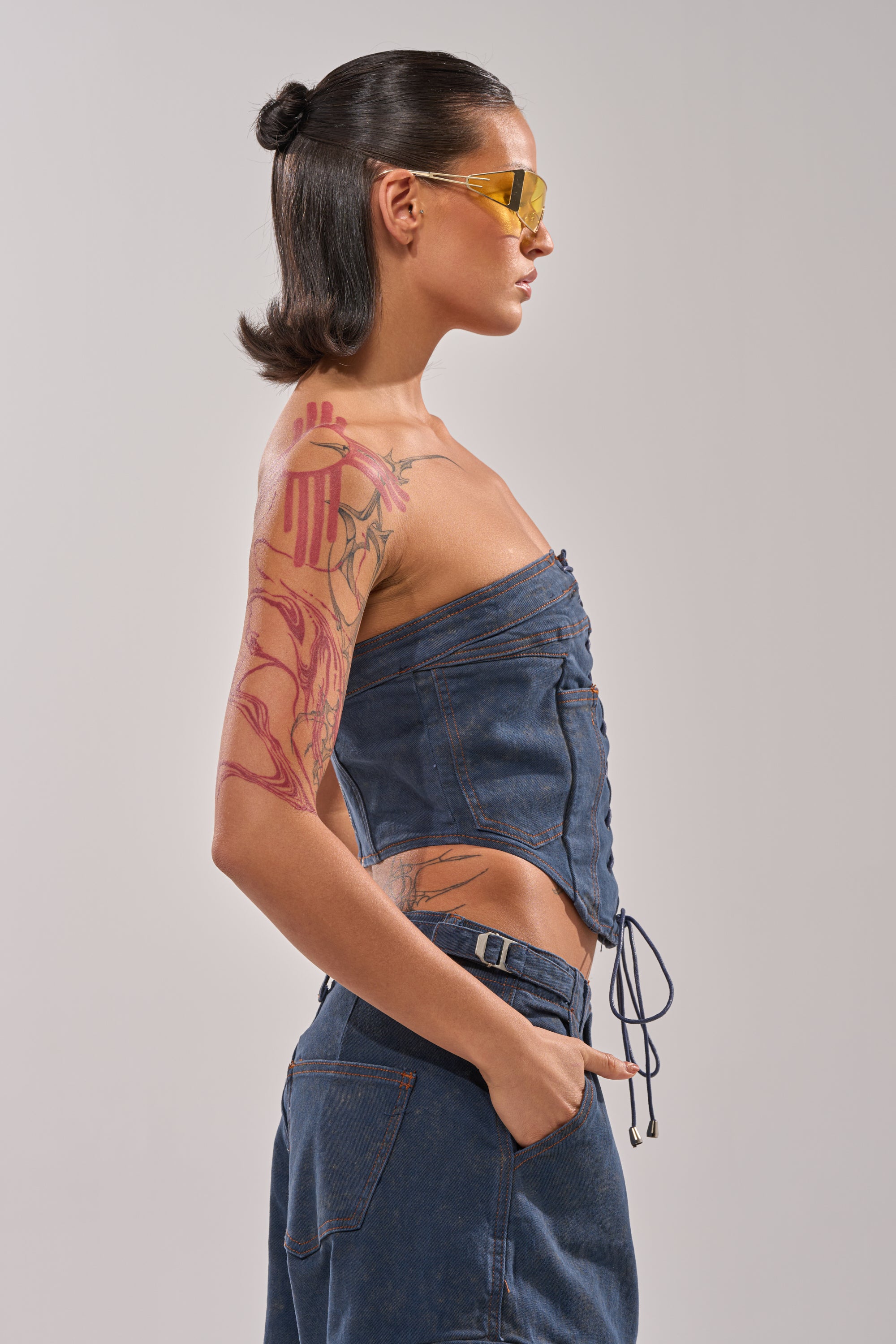 A person with slicked-back hair, yellow sunglasses, and colorful tattoos strikes a profile pose with one hand in their pocket, wearing the BOLD LACE UP CORSET for a vibrant Y2K-inspired look. 2