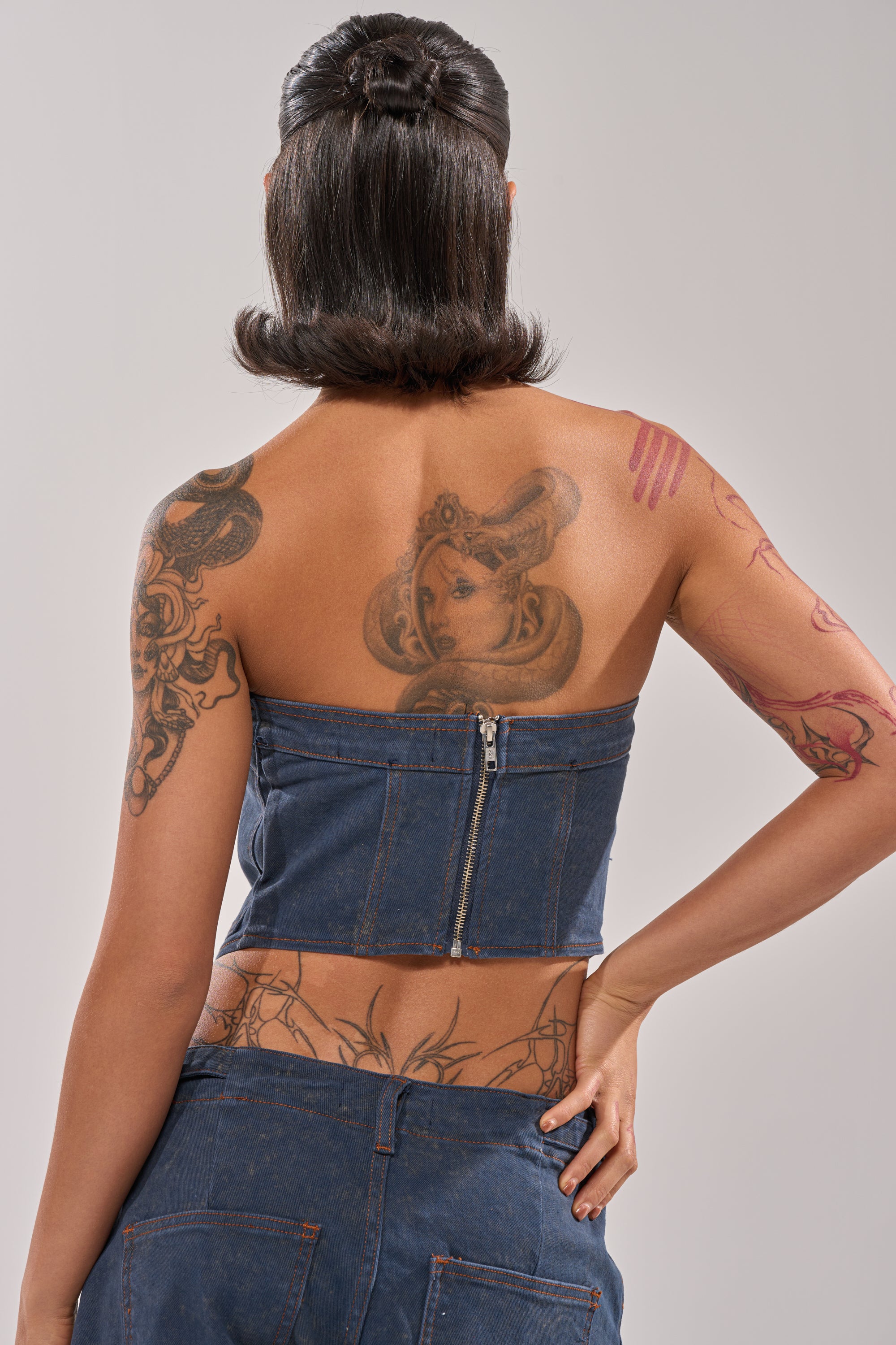A person with shoulder-length dark hair is seen from the back flaunting Y2K vibes in the BOLD LACE UP CORSET and matching pants, highlighting various tattoos on their back and arms. 3