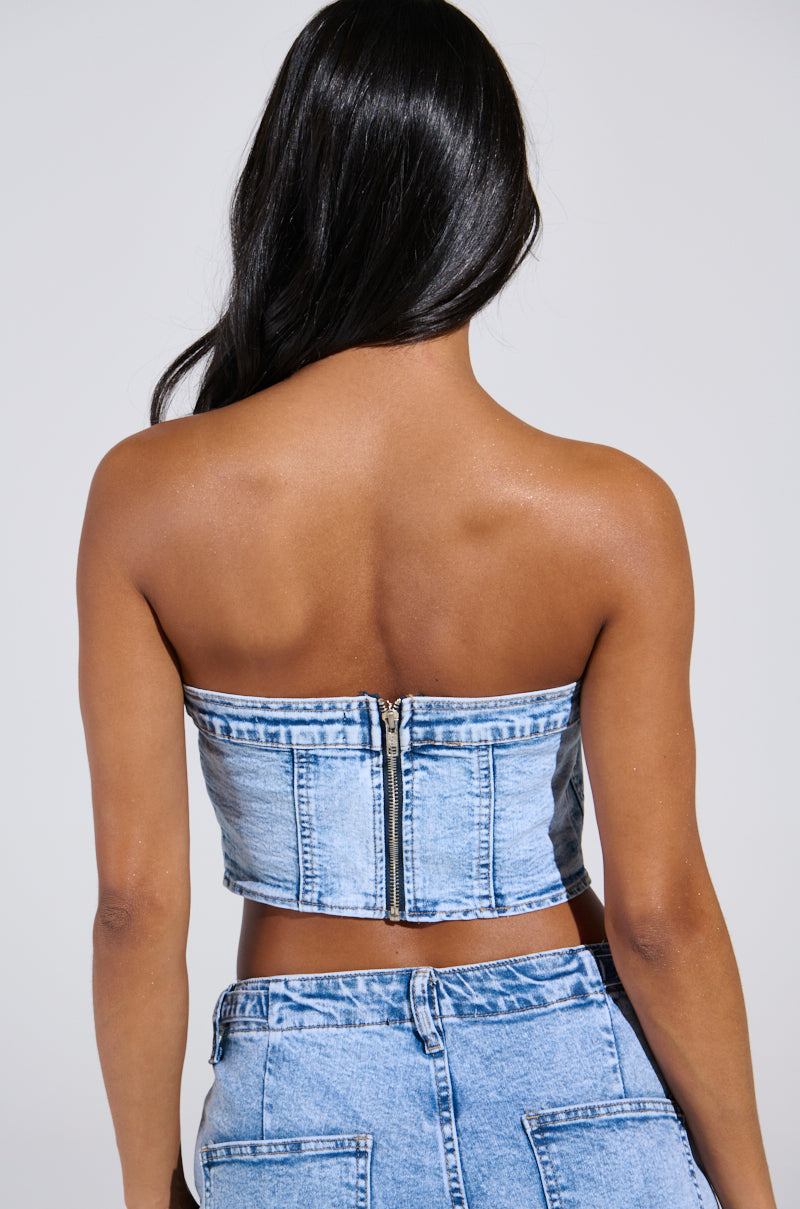 A person with long black hair, facing away, wears the TIED UP DENIM CORSET and matching jeans. The strapless denim top has a visible back zipper resembling a corset. The background is plain and light-colored.