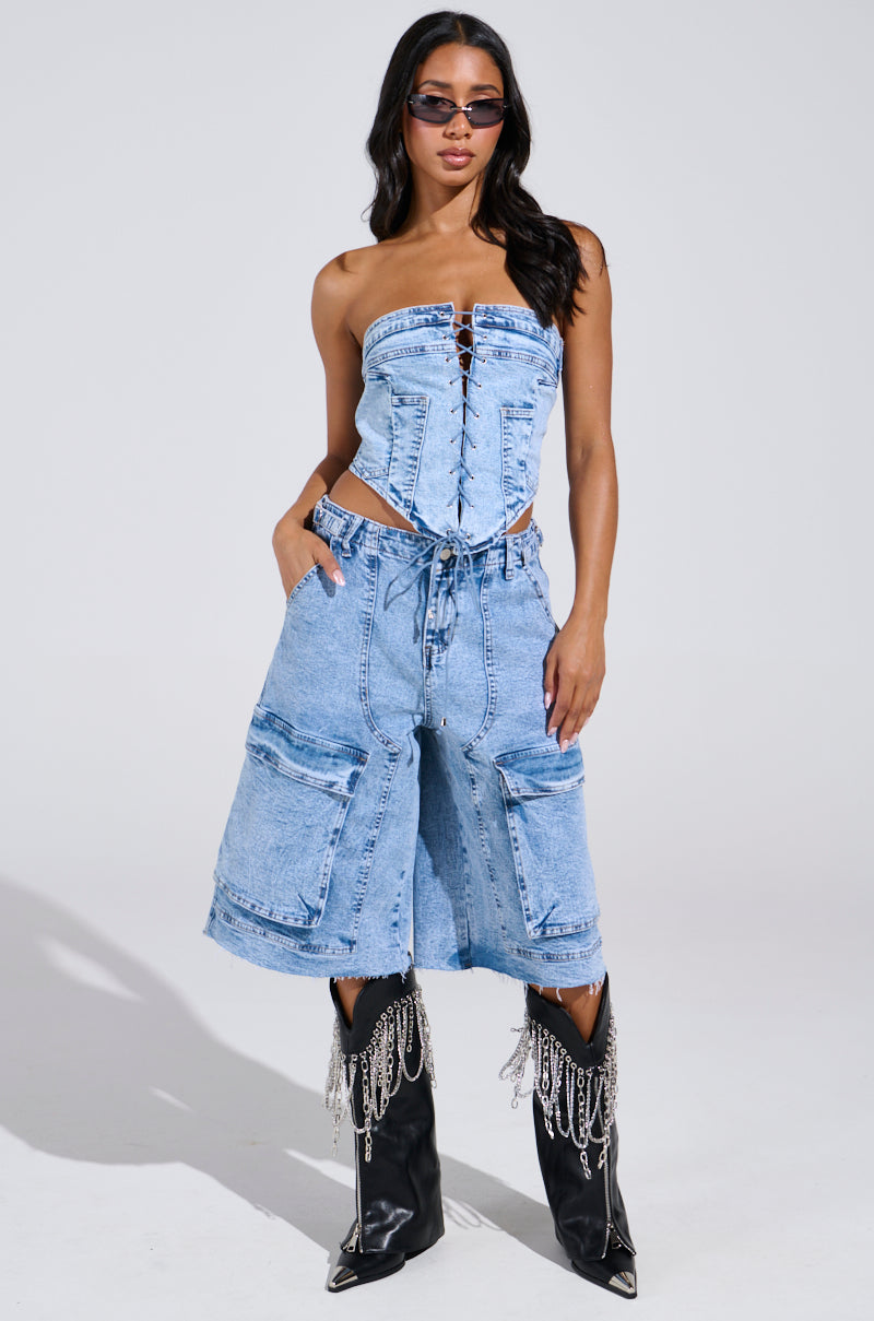 Wearing sunglasses, a person strikes a pose in the TIED UP DENIM CORSET and light blue knee-length shorts with large pockets. They complete their style with knee-high black boots decorated with chains, set against a plain white backdrop.