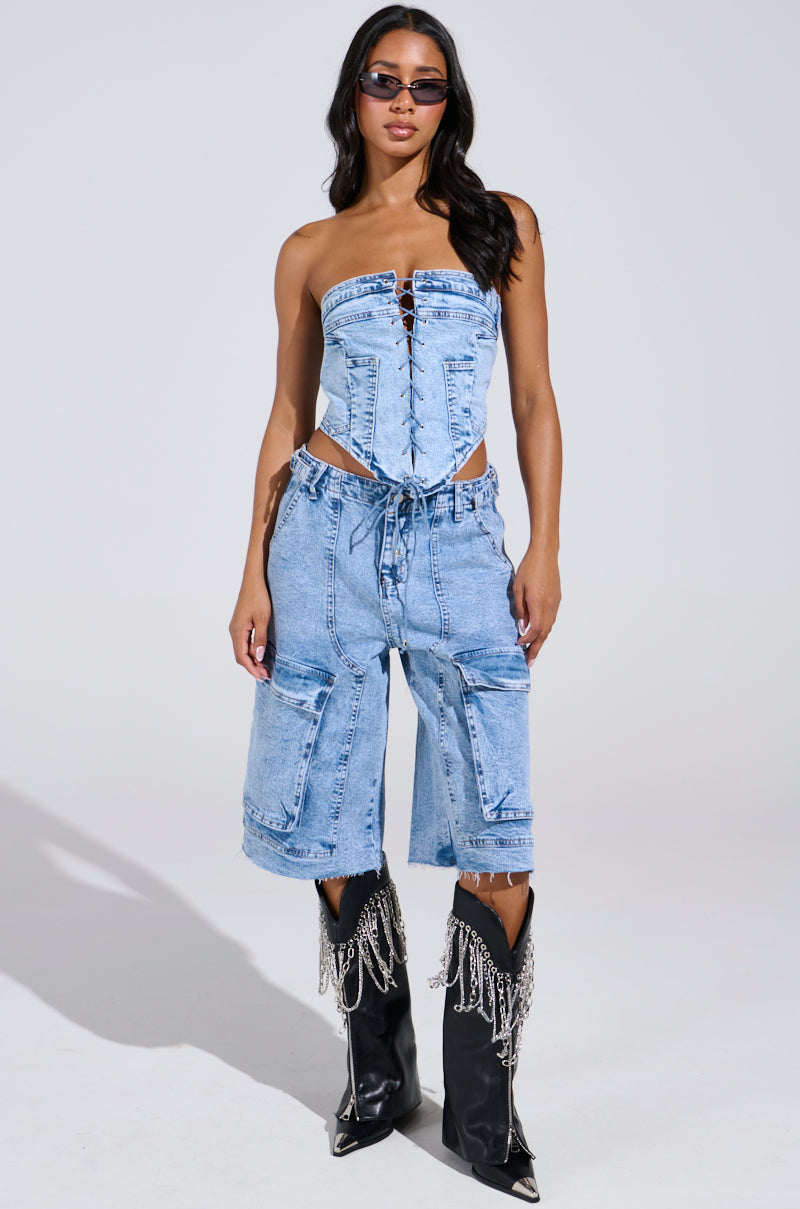 Donning the TIED UP DENIM CORSET with a lace-up front, dark sunglasses, denim capri pants, and black knee-high boots with chain fringe, the individual stands confidently against a plain background.