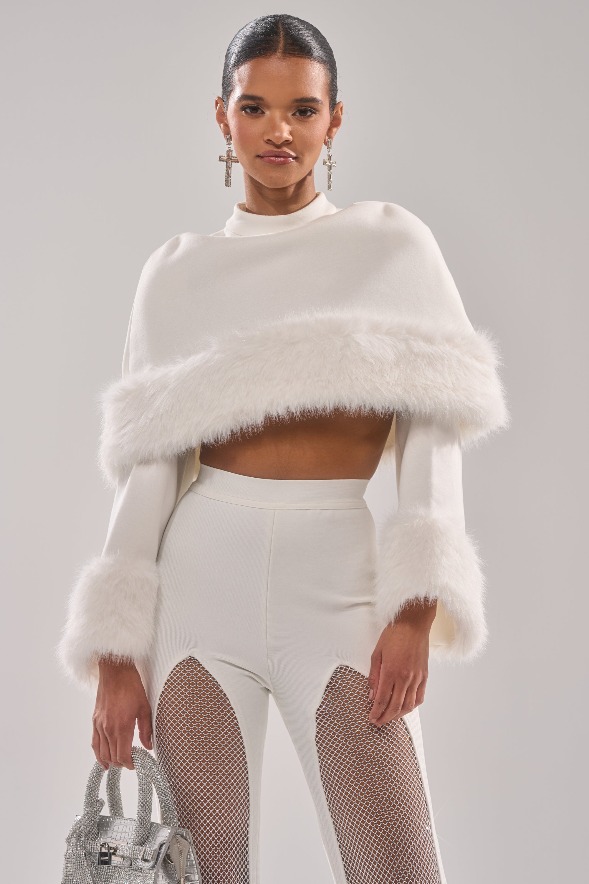 A woman styles the SNOW BUNNY FUR BLOUSE—a white cropped top with faux fur trim—with white mesh-panel pants, a small silver handbag, and cross earrings for a chic winter look.