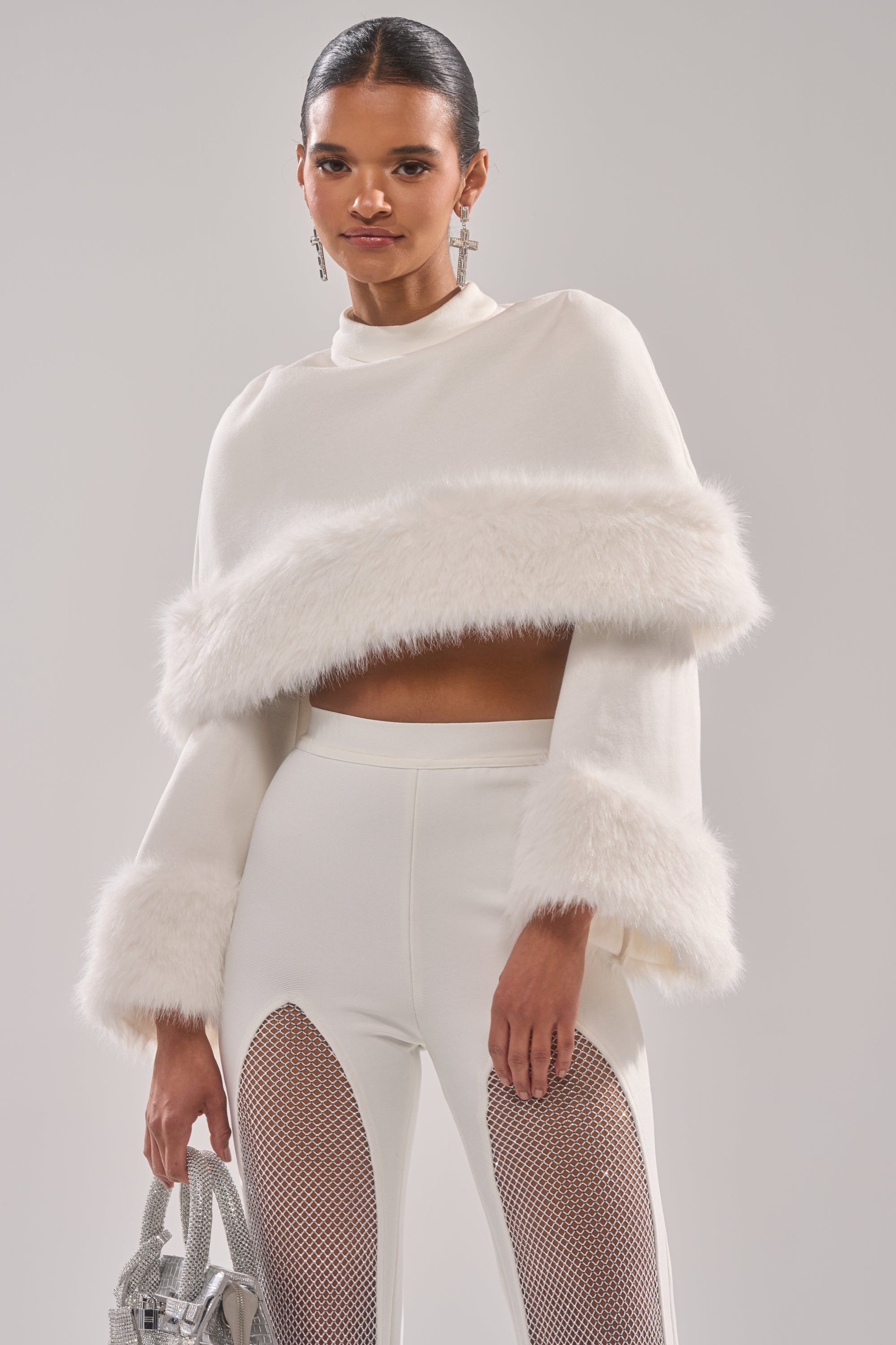 A woman styles the SNOW BUNNY FUR BLOUSE, featuring faux fur trim, with high-waisted white pants with mesh cut-outs, a silver bag, and slicked-back hair for a chic holiday look.