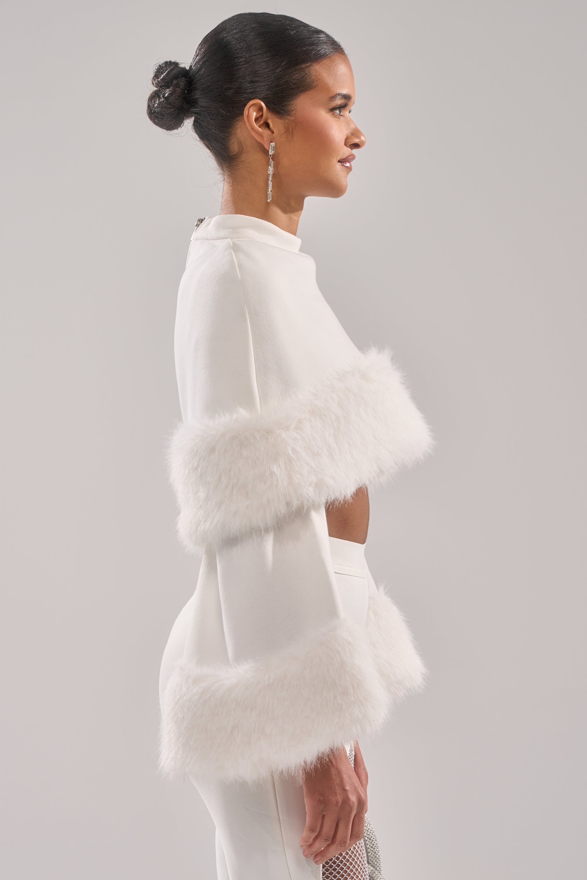 A woman in profile models the SNOW BUNNY FUR BLOUSE, featuring wide sleeves and a fluffy fur-trimmed hem. She wears her hair in a neat bun with dangling earrings, creating a chic holiday look against a plain gray background.