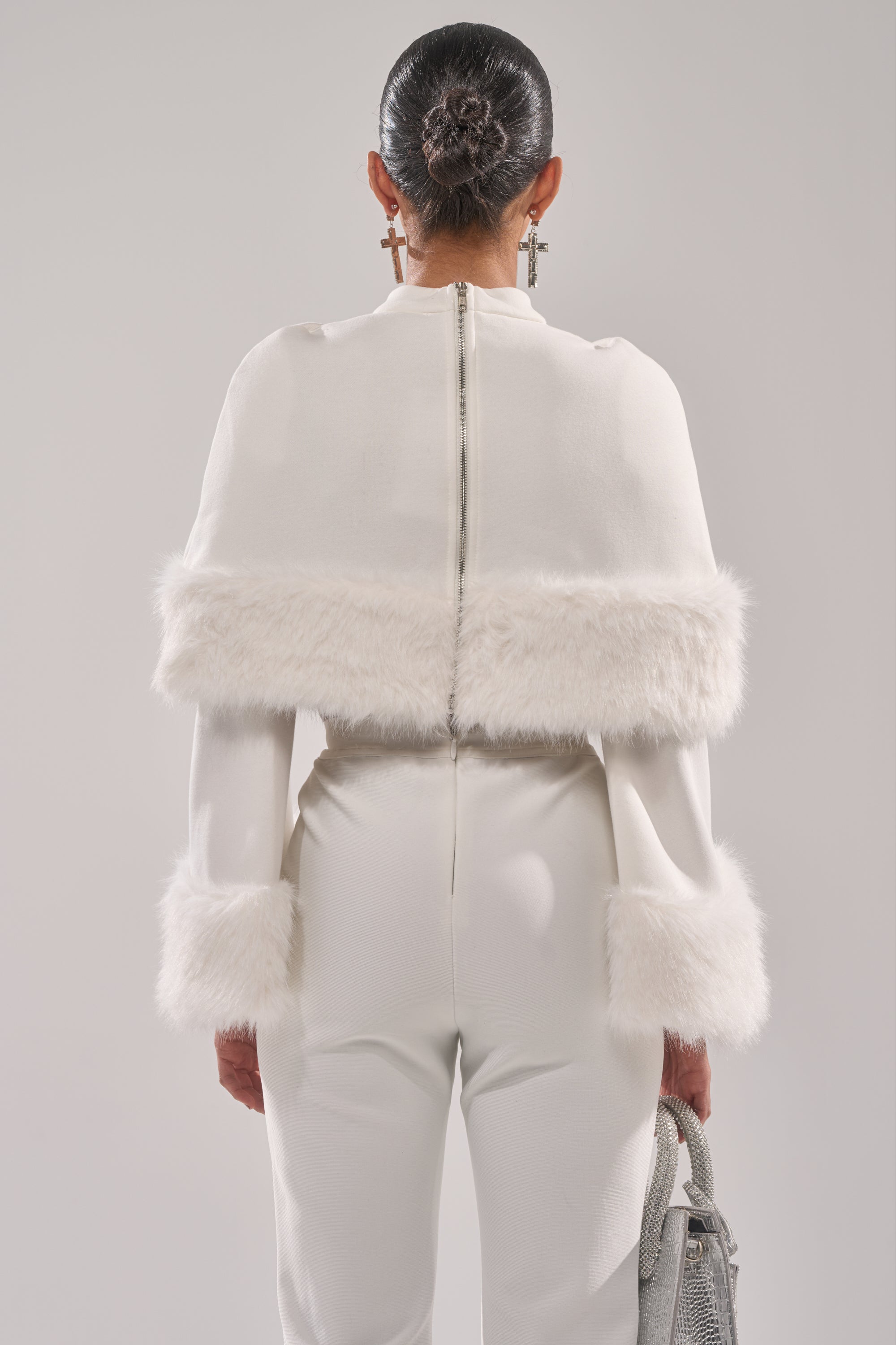 A woman faces away, wearing the SNOW BUNNY FUR BLOUSE featuring fur accents on the sleeves and shoulders. Her hair is in a sleek bun, and she carries a silver textured handbag—ideal for chic winter style.