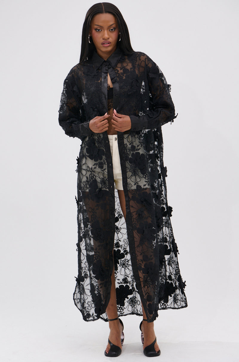 A woman faces forward wearing the AMARIA LONG LACE BUTTON UP SHIRT in black over a black bra and white shorts, styled with black heeled sandals against a plain white background.