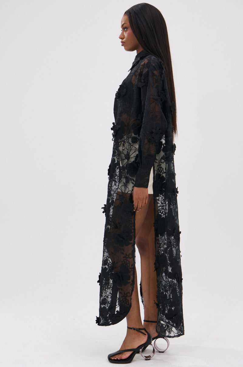 A woman stands in profile against a plain white background, wearing the AMARIA LONG LACE BUTTON UP SHIRT as a dress, styled with black strappy heels. She has long, straight hair and the lace shirt features sheer details and a high slit.