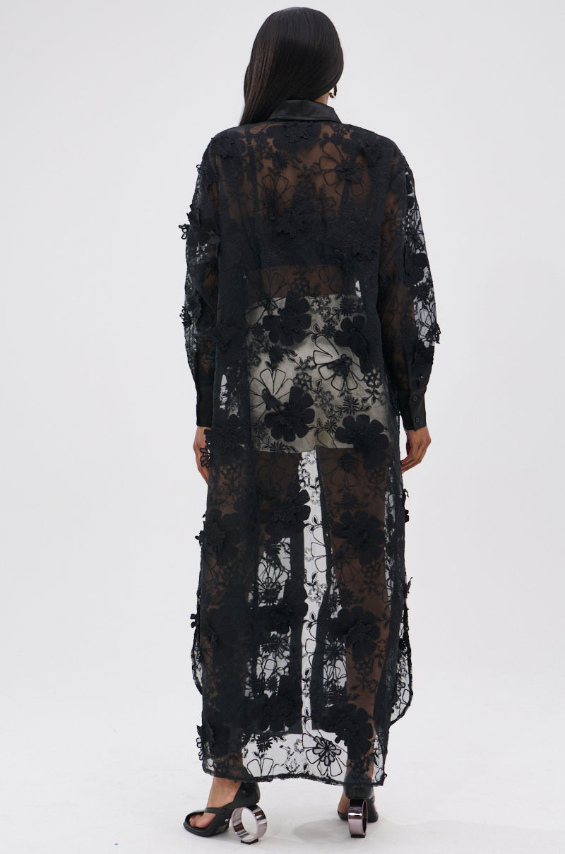 A person stands facing away against a plain white background, wearing the AMARIA LONG LACE BUTTON UP SHIRT in black over matching lace shorts, showcasing its sheer floral pattern and lightweight design.