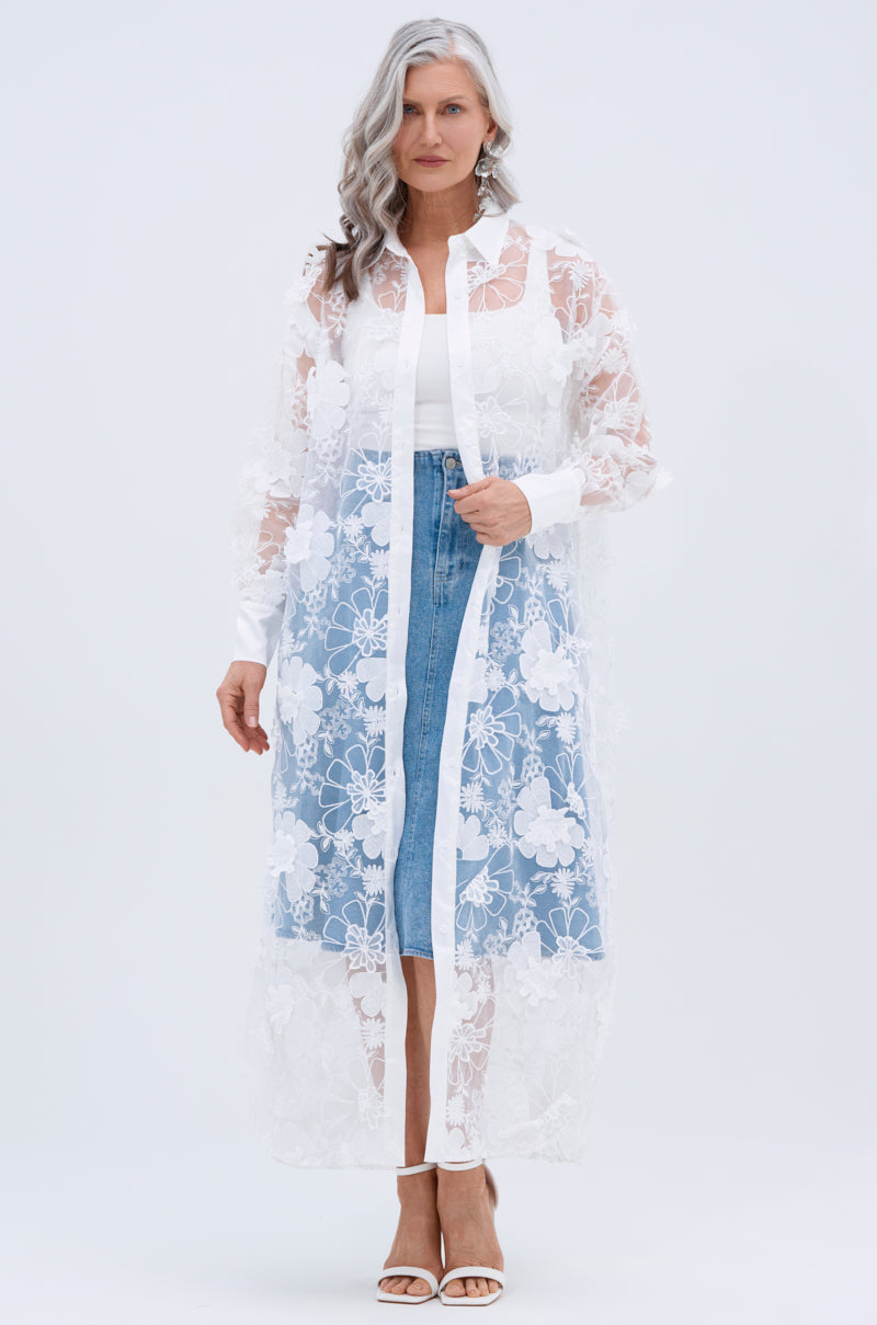 A woman with long, wavy gray hair wears the LACE BUTTON UP—a sheer mesh blouse with floral appliques—over a white top and blue denim skirt. She stands facing forward in white sandals against a plain light background.