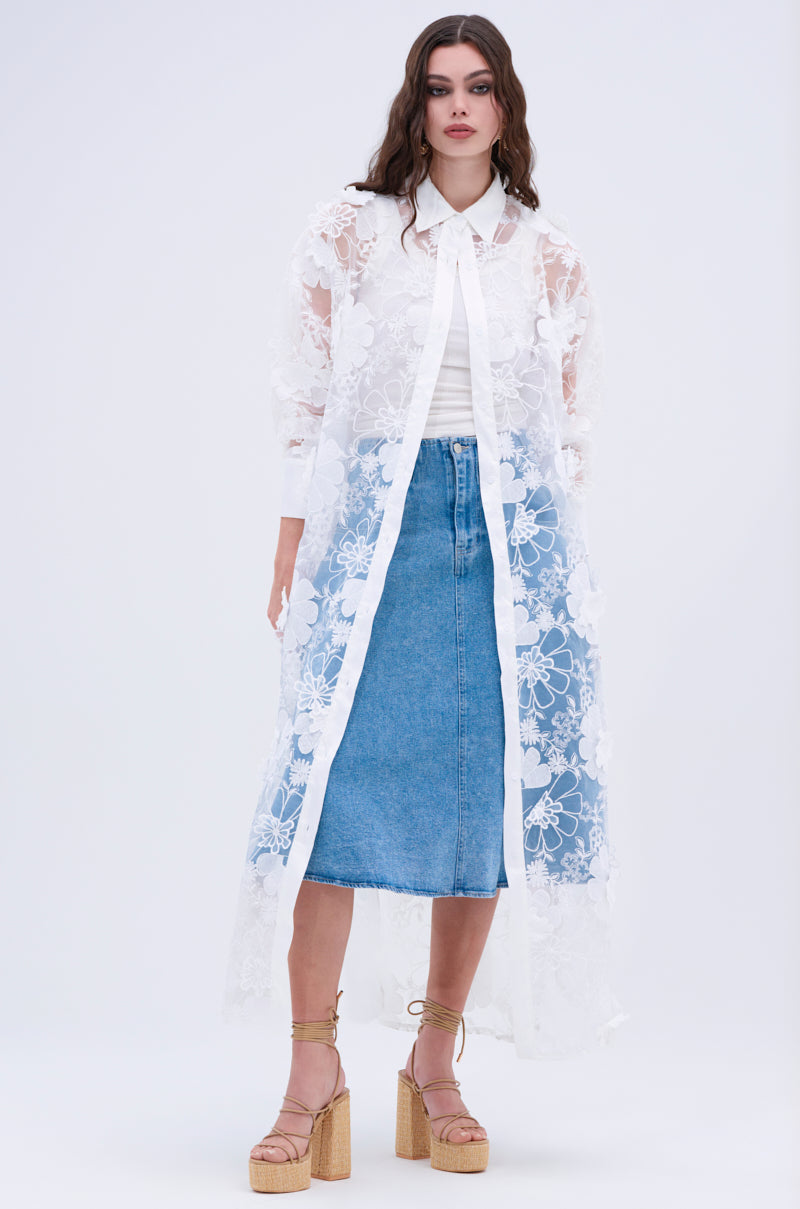 A woman stands against a plain light background, wearing the LACE BUTTON UP—a sheer, long white lace cardigan—over a white blouse and blue denim midi skirt, finished with beige platform sandals.
