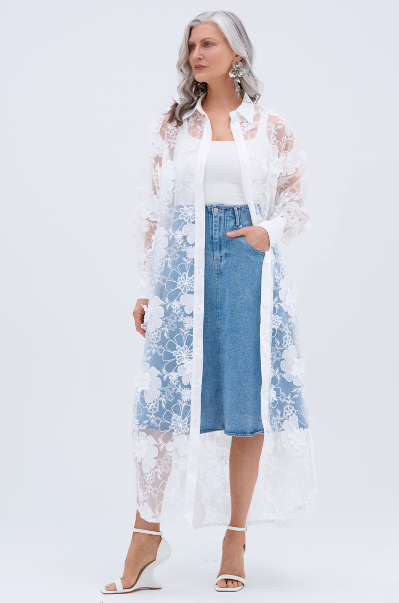A woman with long gray hair wears the LACE BUTTON UP sheer mesh blouse with floral appliques over a white top and a knee-length blue denim skirt, paired with white heeled sandals, standing against a plain light background.