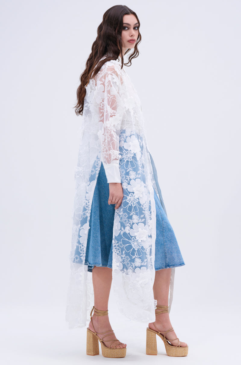A woman with long wavy hair wears the LACE BUTTON UP sheer mesh blouse with floral appliques over a blue denim dress, styled with gold platform sandals, posing against a plain light background.