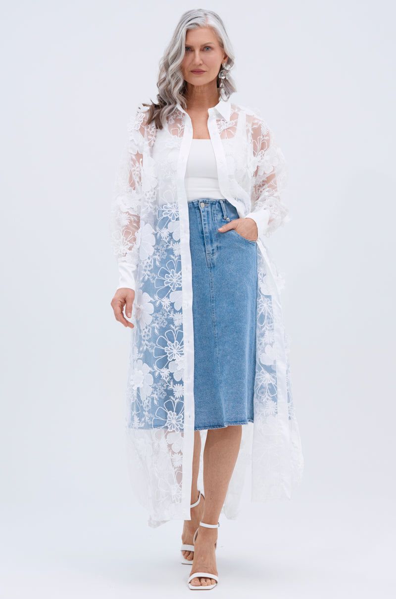 A woman with long gray hair models the LACE BUTTON UP worn as a sheer duster over a white top and knee-length denim skirt, paired with white heeled sandals, against a plain light background.