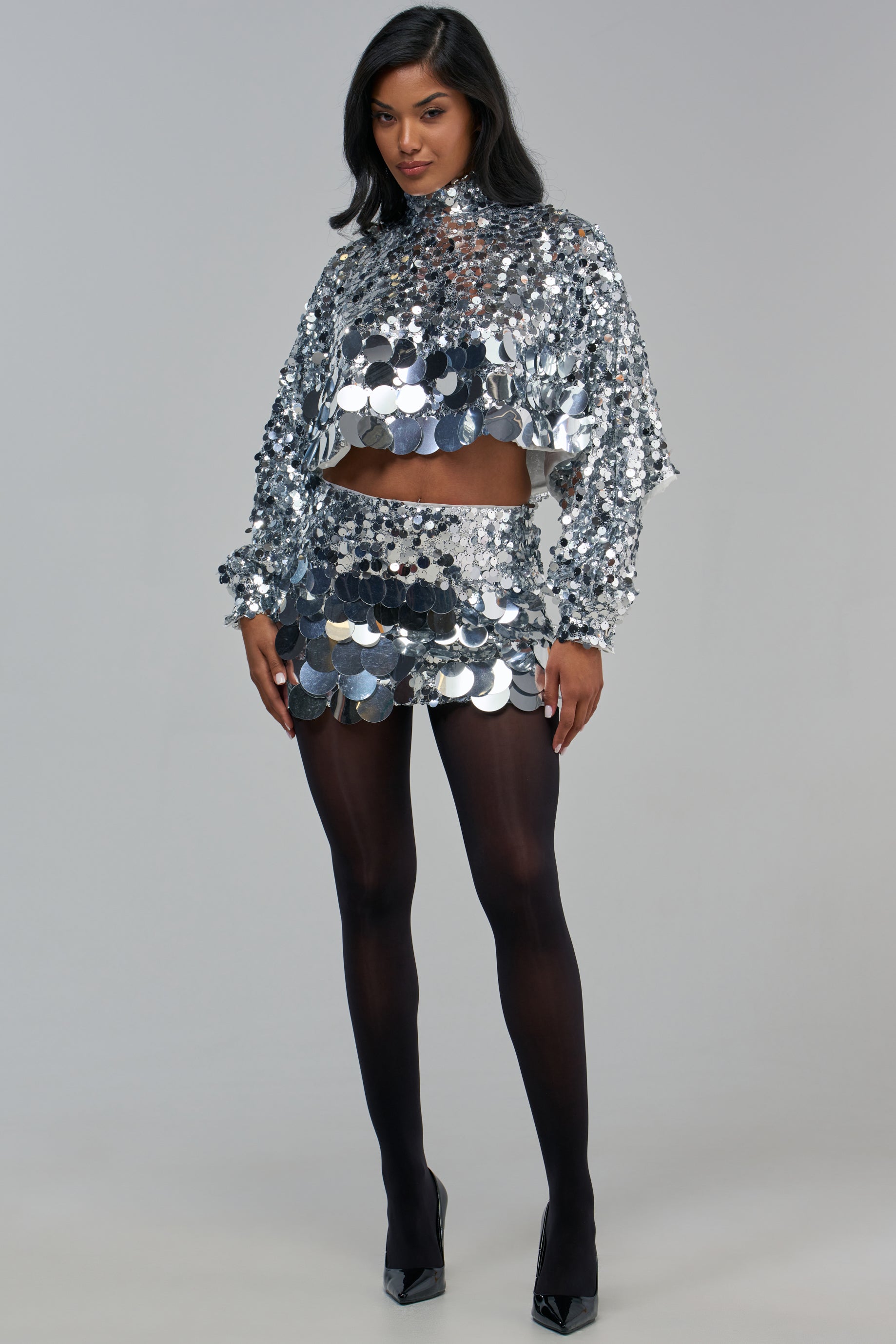 A person wearing a long-sleeved silver sequin crop top and a matching HOLIDAY BLING SEQUIN MINI SKIRT with black tights and black heels.