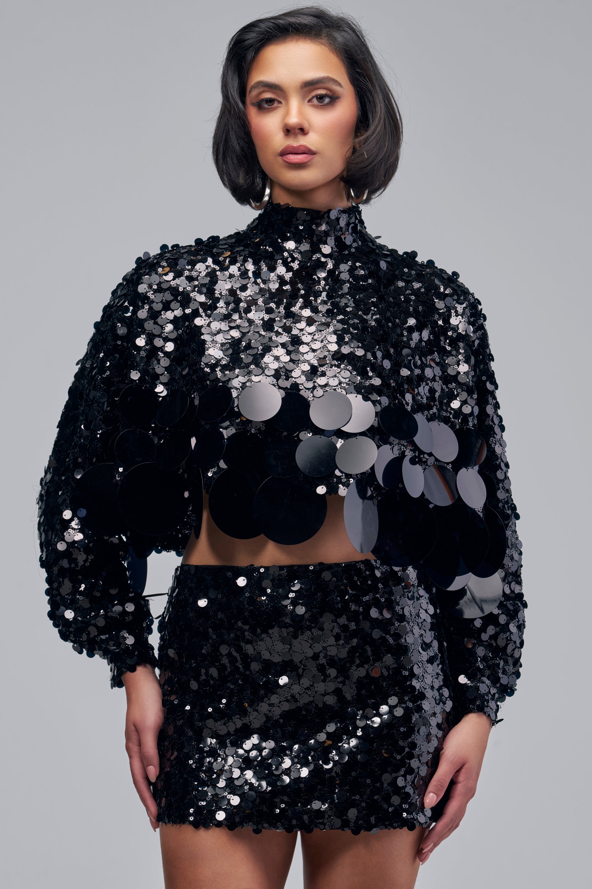 A person wears the HOLIDAY BLING SEQUIN BLOUSE IN BLACK paired with a high-waisted skirt covered in large, circular, reflective sequins.