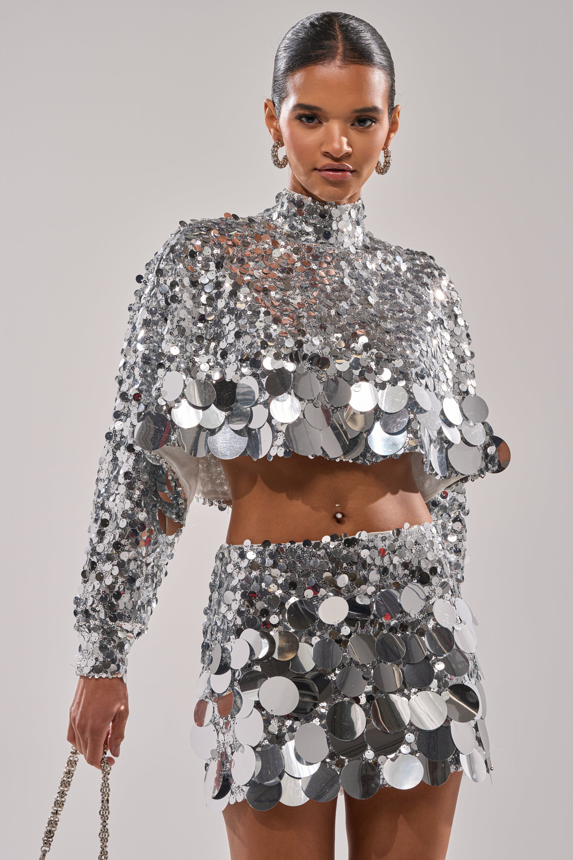 Wearing the HOLIDAY BLING SEQUIN BLOUSE and a matching mini skirt with metallic details, a woman poses confidently against a plain background, her hair slicked back and hoop earrings completing the look.