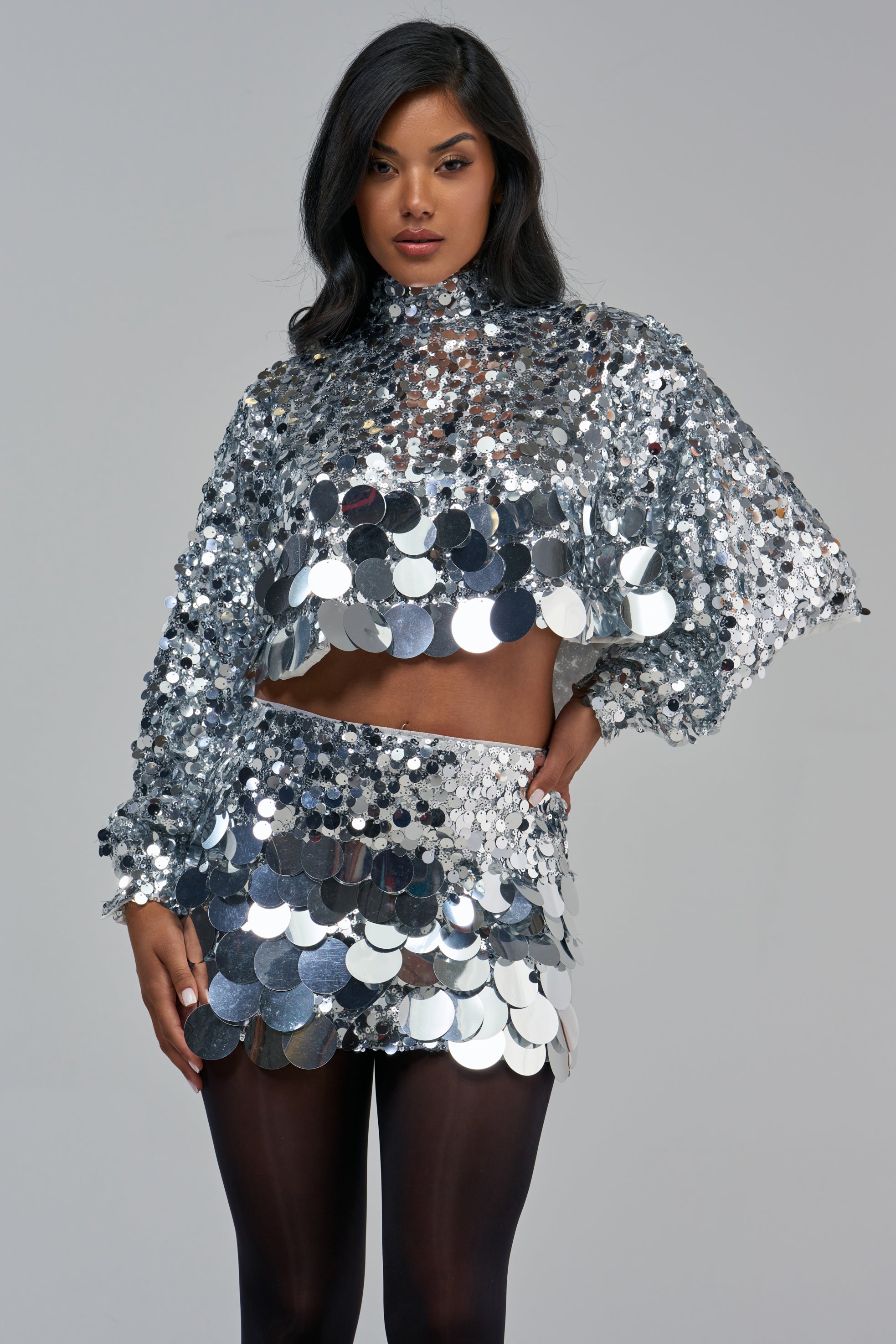 A person wears a HOLIDAY BLING SEQUIN BLOUSE and a matching mini skirt, both covered in large, reflective silver sequins.