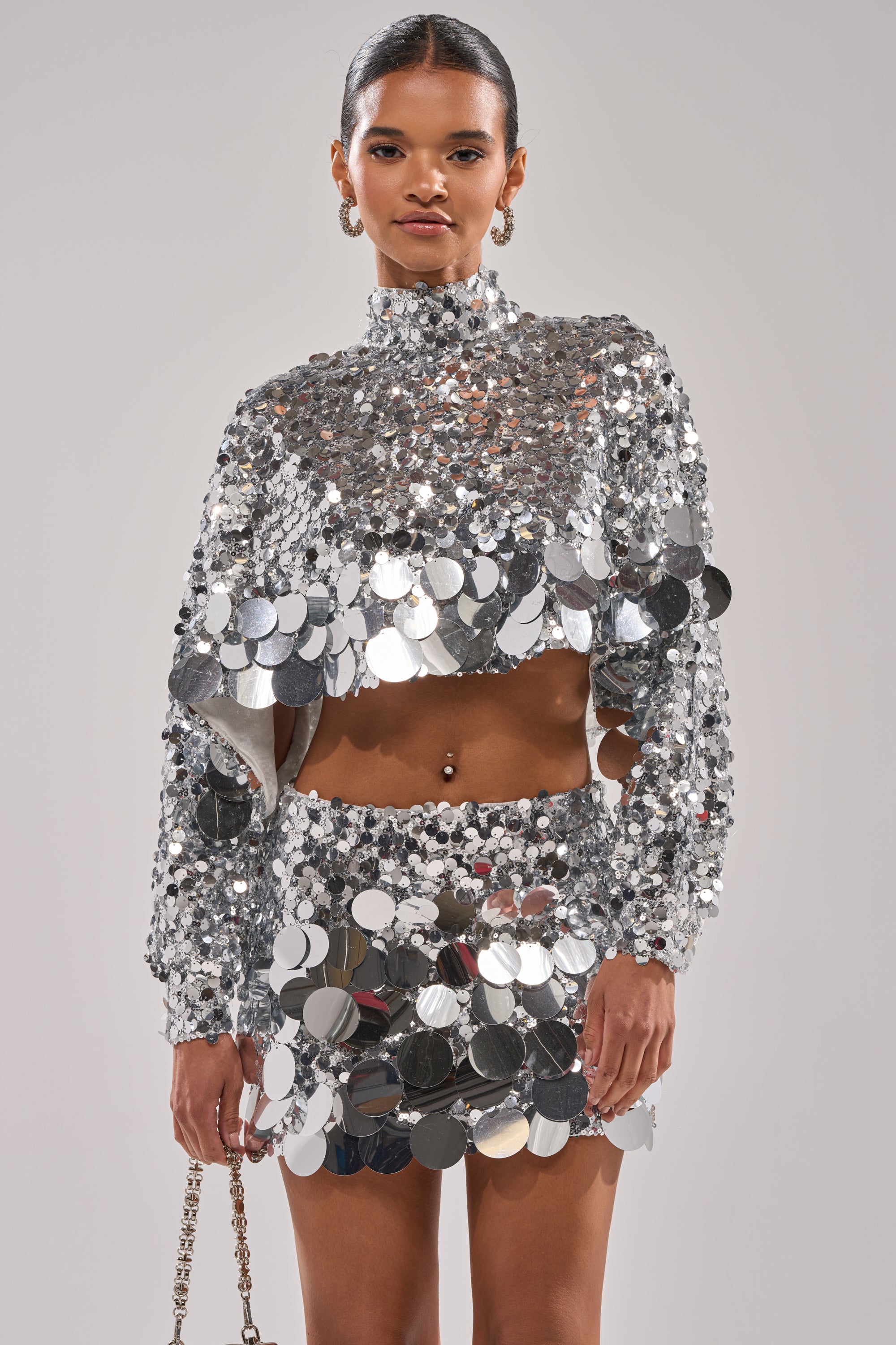 A woman poses confidently against a plain background, wearing the HOLIDAY BLING SEQUIN BLOUSE crop top and matching mini skirt with large shiny silver discs. She accessorizes with a chain-strap handbag and slicked-back hair.