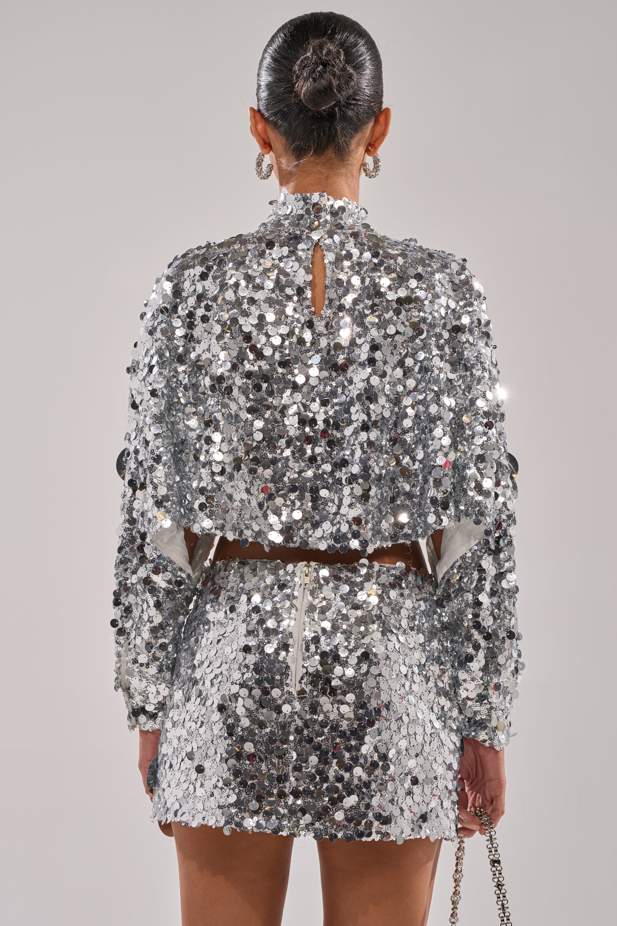 A person is seen from the back wearing the HOLIDAY BLING SEQUIN BLOUSE and a matching mini skirt with metallic embellishments. Their hair is in a sleek bun, and they carry a small chain strap bag.