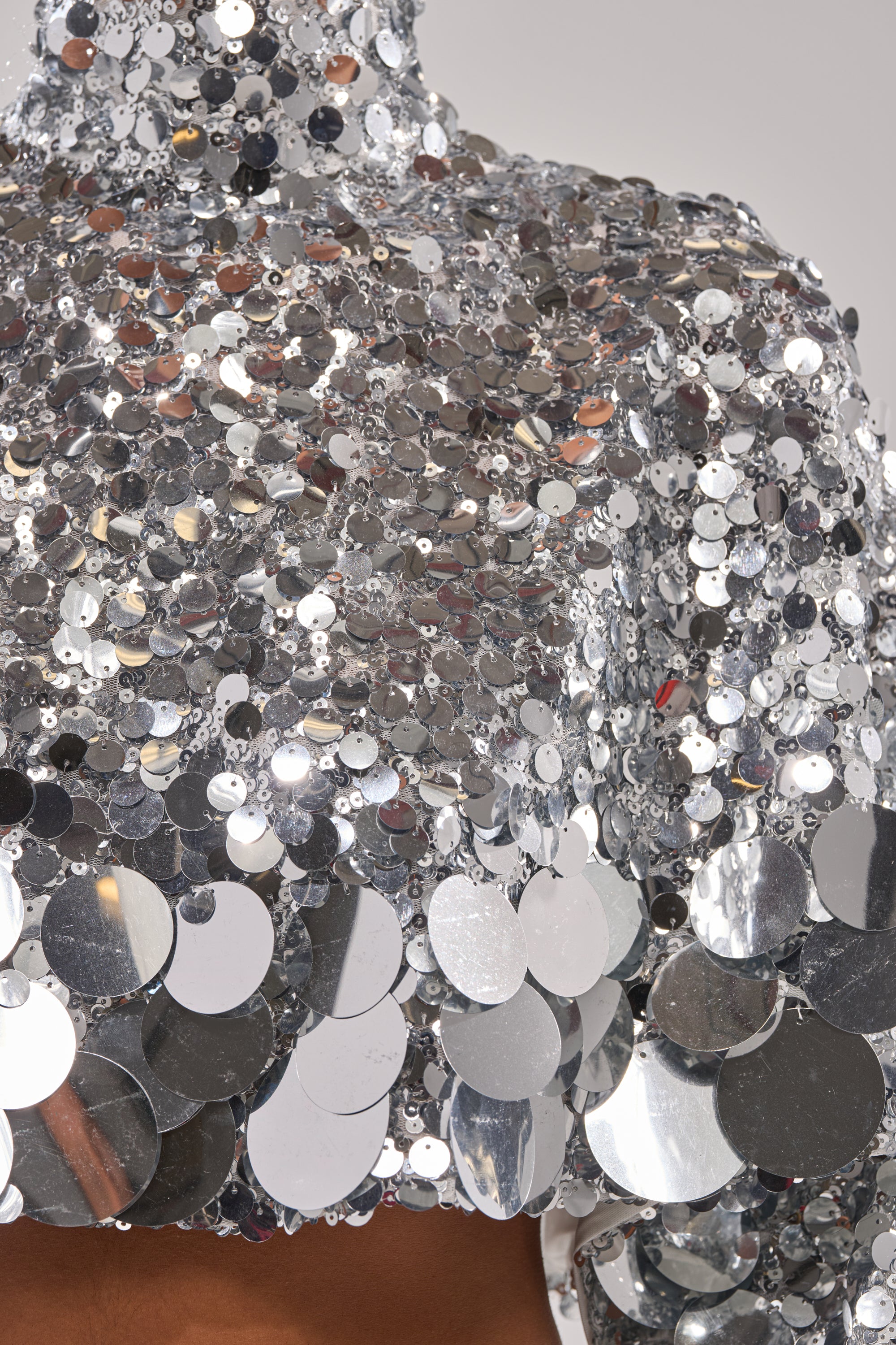 Close-up of the HOLIDAY BLING SEQUIN BLOUSE featuring oversized shiny silver sequins that reflect light, creating a textured, metallic look. A glimpse of skin is visible at the hem against a neutral background.