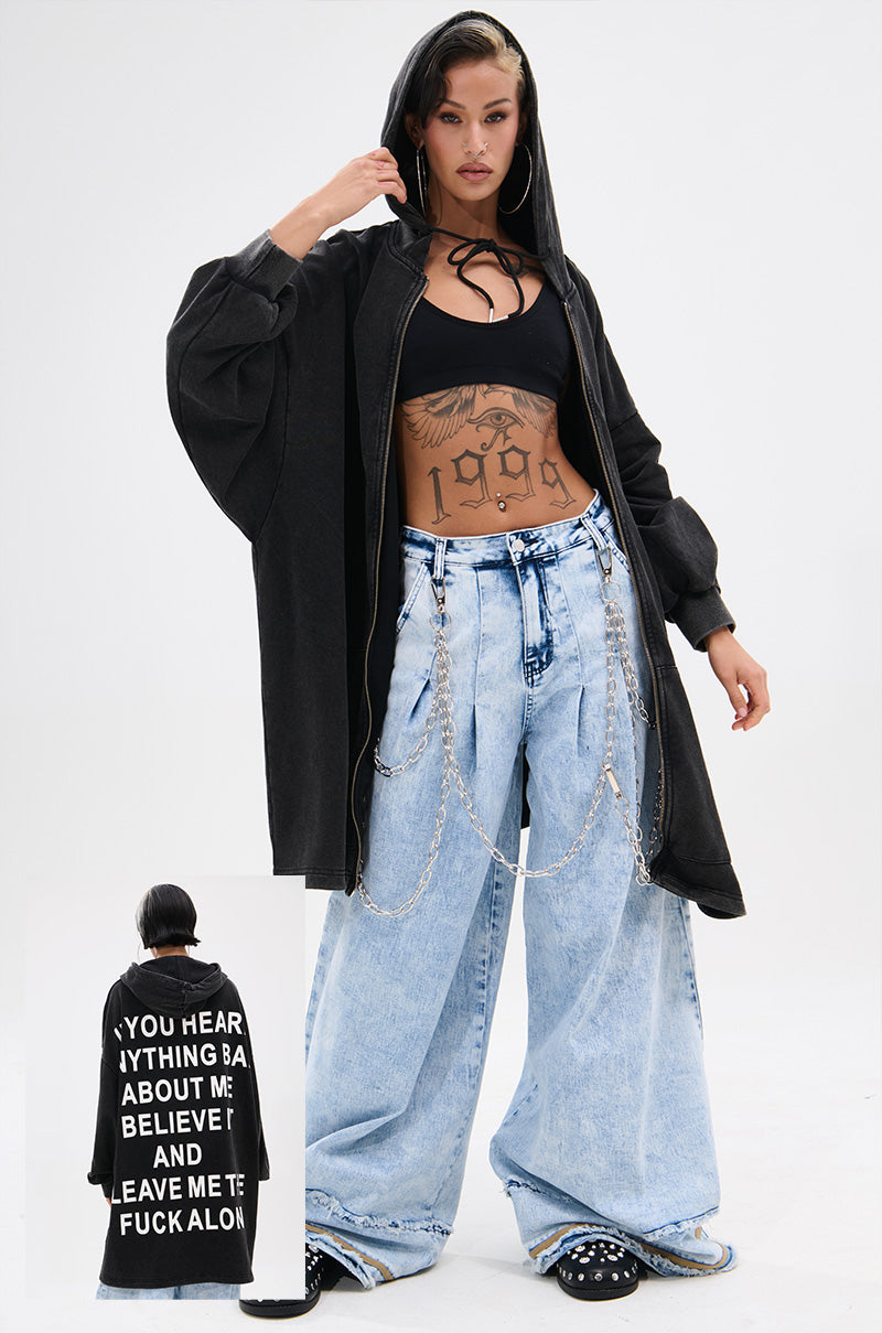 A woman poses confidently in the BELIEVE WHAT YOU HEARD OVERSIZED ZIP UP SWEATSHIRT, featuring a relaxed fit and hood. She pairs it with a crop top and wide-leg jeans with chains, accessorized with hoop earrings, tattoos, and dark lipstick. 0