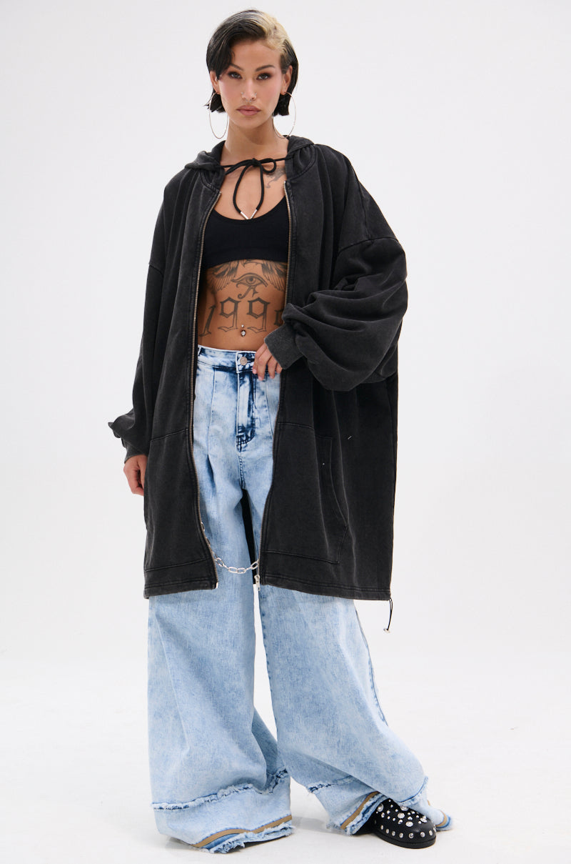 A person with short dark hair and visible torso tattoos wears a black crop top, the BELIEVE WHAT YOU HEARD OVERSIZED ZIP UP SWEATSHIRT, wide-leg light blue jeans with chain detail, and black sneakers with white accents. 9