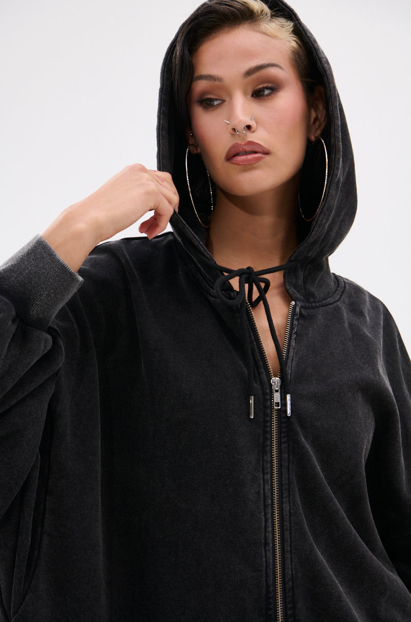 Wearing the BELIEVE WHAT YOU HEARD OVERSIZED ZIP UP SWEATSHIRT with the hood up, a person with hoop earrings and nose piercings looks serious, gazing to the side and raising one hand near their face. 12