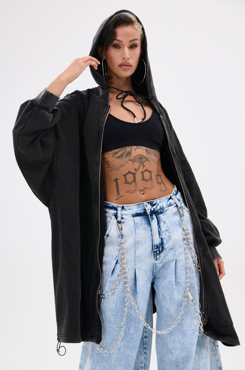 A woman poses confidently in the BELIEVE WHAT YOU HEARD OVERSIZED ZIP UP SWEATSHIRT, styled with light blue jeans, chain accessories, a black crop top, large hoop earrings, visible torso tattoos, and one hand holding her hood. 1