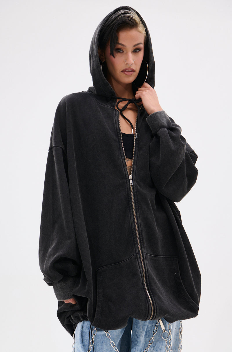 Model wears the BELIEVE WHAT YOU HEARD OVERSIZED ZIP UP SWEATSHIRT with hood up over a black top and pairs it with light blue jeans featuring silver chains, standing against a plain white background. 2
