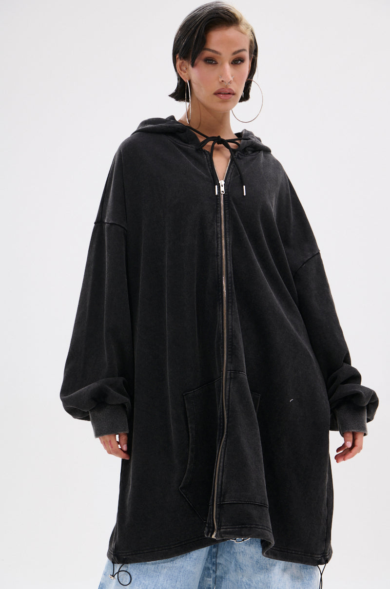 A person with short dark hair and large hoop earrings wears the BELIEVE WHAT YOU HEARD OVERSIZED ZIP UP SWEATSHIRT in black, featuring a relaxed fit, front pocket, and fleece-like fabric, styled with light blue jeans against a plain white background. 4