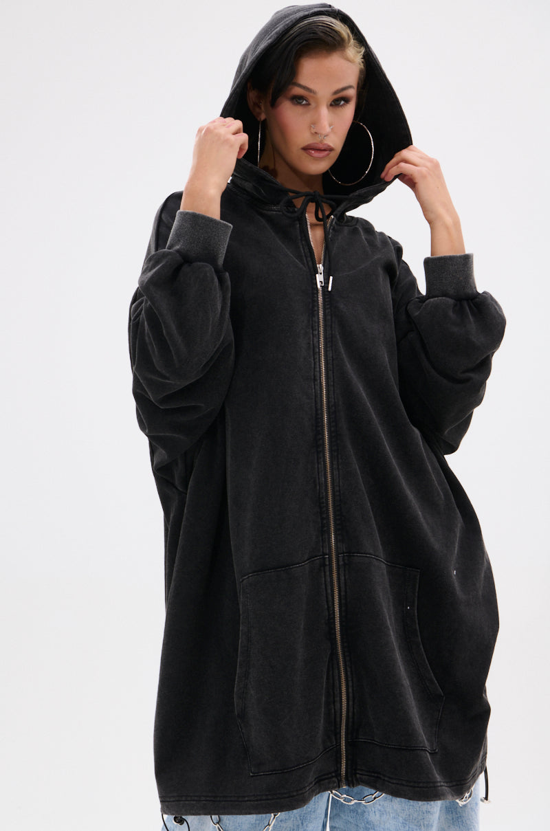 A woman poses against a white background, wearing the BELIEVE WHAT YOU HEARD OVERSIZED ZIP UP SWEATSHIRT—a loose black fleece zip-up hoodie with a hood up, oversized sleeves, front pockets, and a relaxed silhouette. 5