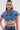 A woman wears the FULL OF LIFE DENIM BUTTON DOWN WITH PLAID DETAILS, a boxy cropped denim shirt, paired with high-waisted blue jeans. She accessorizes with hoop earrings and slicked-back hair.