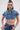A woman wears the FULL OF LIFE DENIM BUTTON DOWN WITH PLAID DETAILS, a boxy cropped silhouette with frayed edges, paired with high-waisted denim pants. She accessorizes with hoop earrings and has slicked-back hair and a confident look.