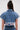 A woman with long dark hair in a bun faces away, wearing the FULL OF LIFE DENIM BUTTON DOWN WITH PLAID DETAILS— a boxy, cropped short-sleeve jacket with frayed edges, multicolored plaid hem—paired with high-waisted jeans.