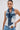 A woman with dark hair tied back wears the ALWAYS RIGHT DENIM ABSTRACT BLOUSE, a patchwork mixed-wash corset-style denim vest, paired with light-wash jeans and hoop earrings, standing against a plain, light background.