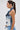 A woman with long brown hair in a ponytail, large hoop earrings, and light blue jeans models the ALWAYS RIGHT DENIM ABSTRACT BLOUSE in profile against a light gray background.